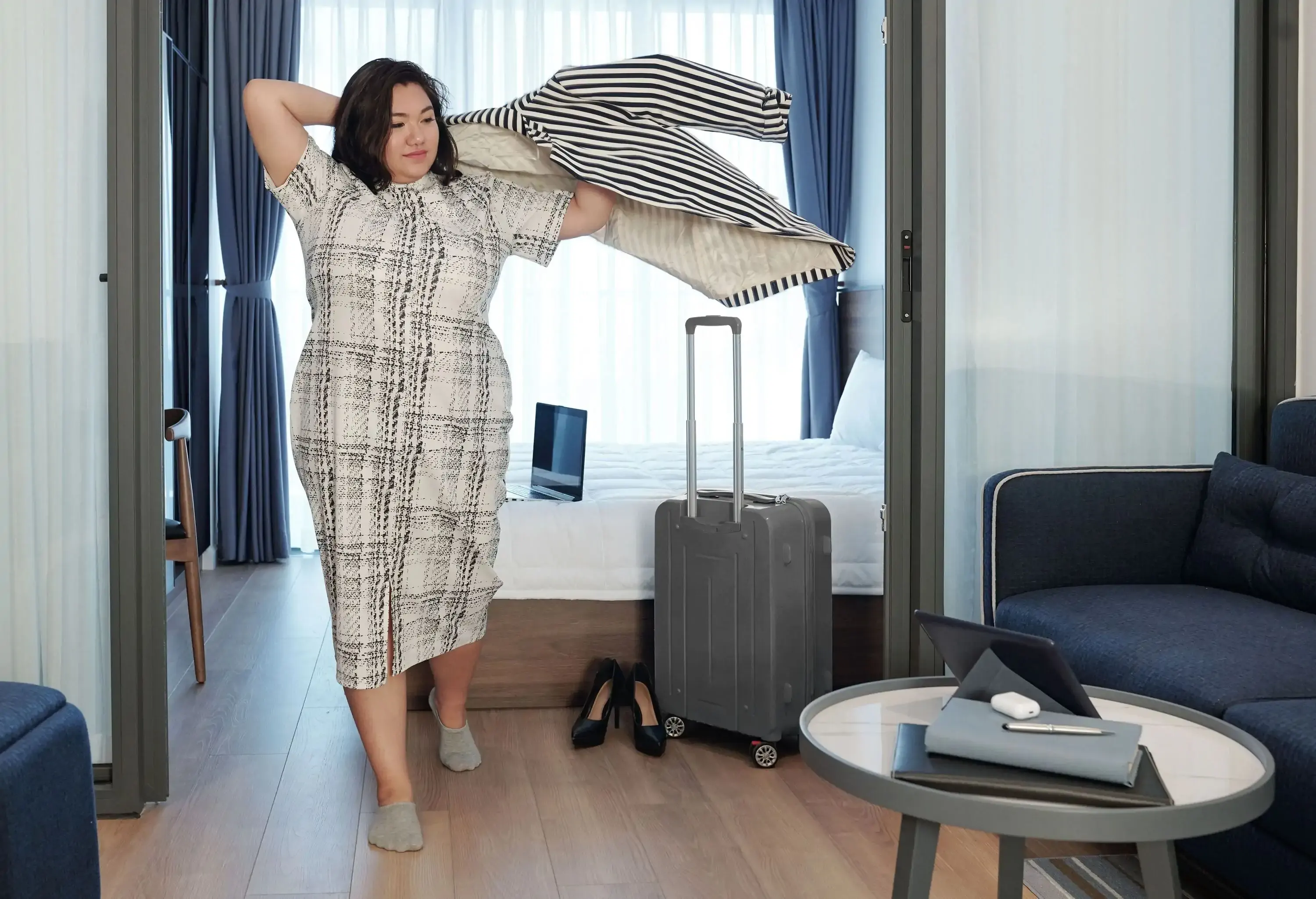A woman putting on an overcoat with her luggage standing by the bed in her hotel room.