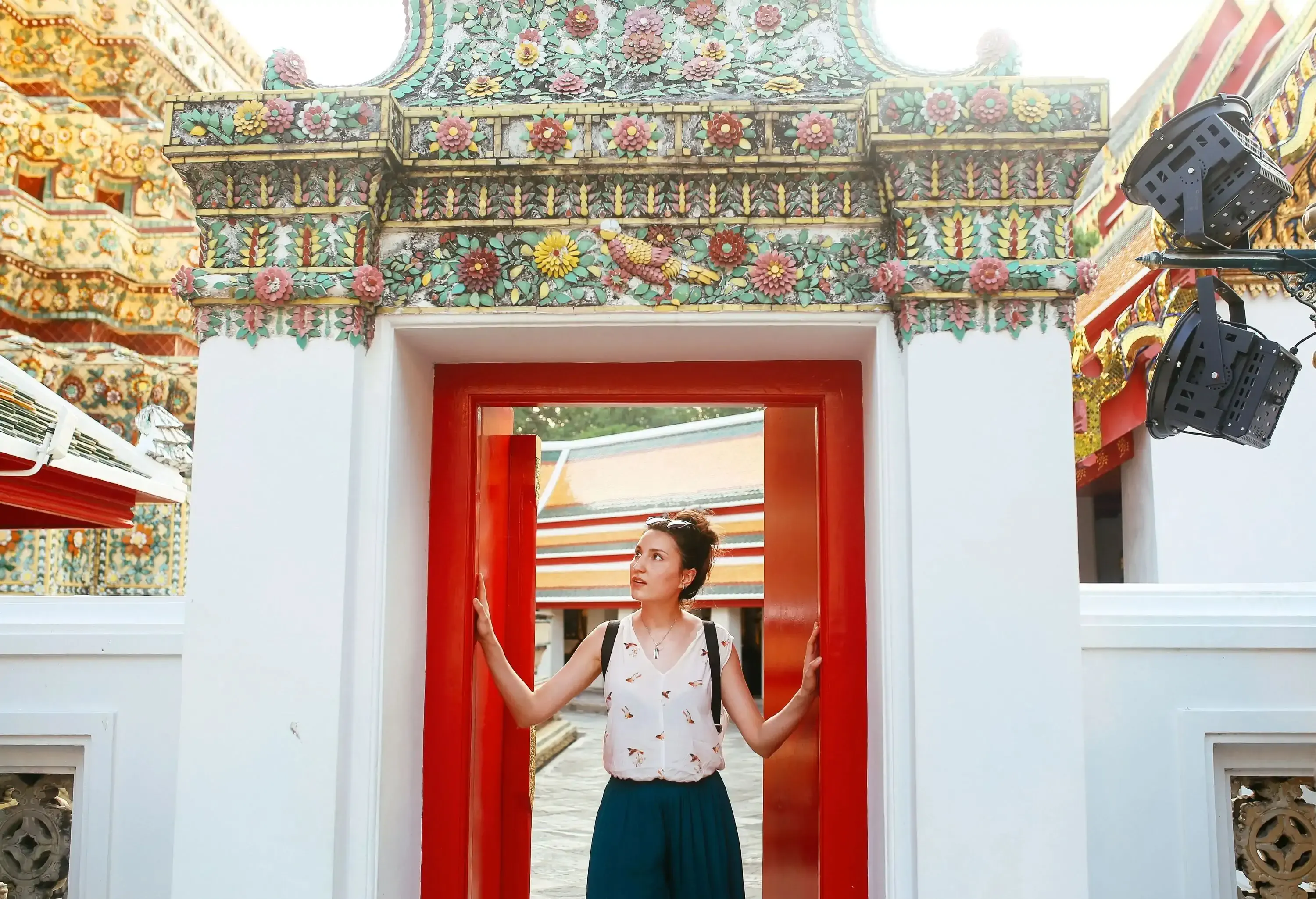 A tourist in casual clothes looks up as she touches the red doors of an entrance with ornate roof design.