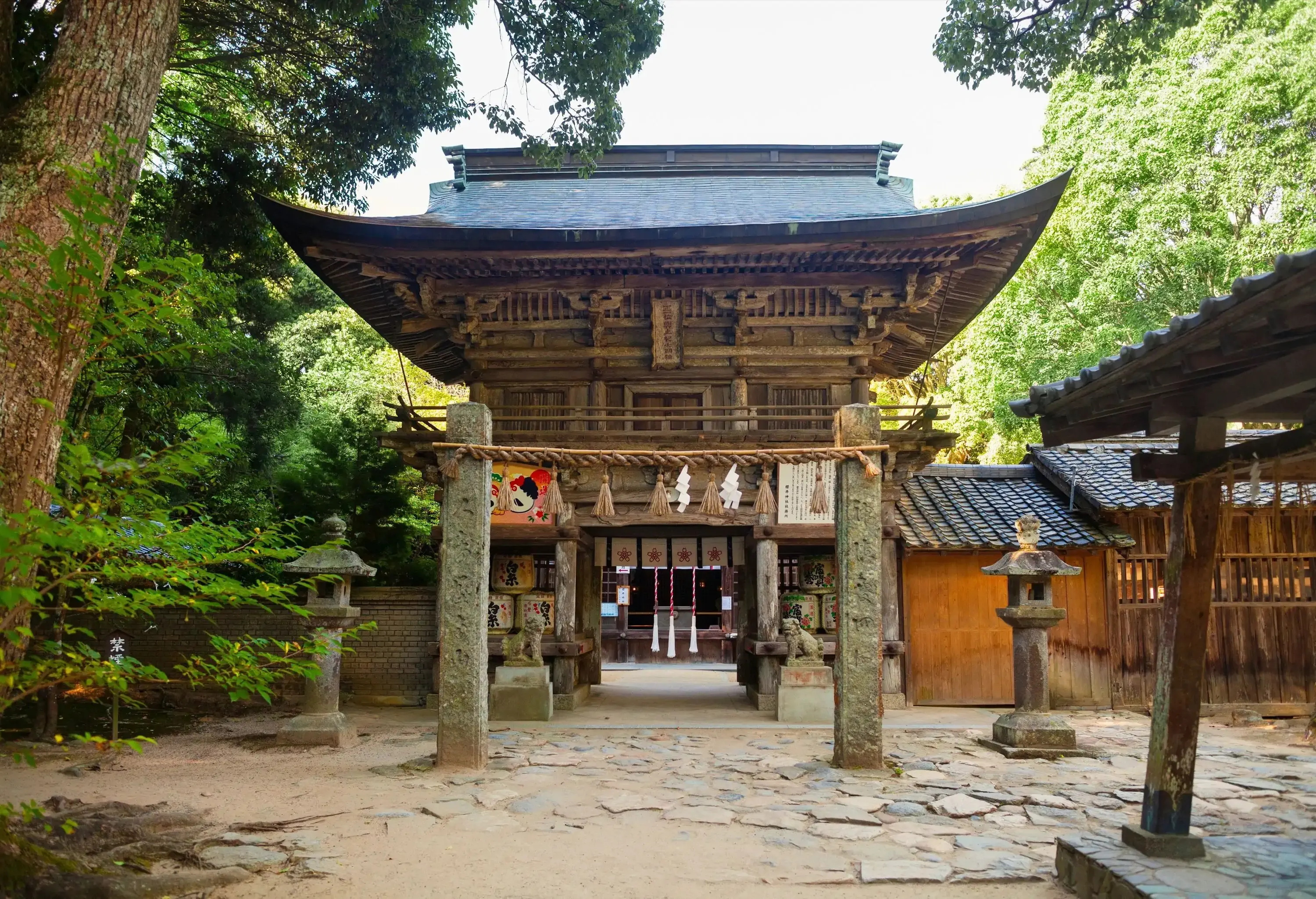 A pathway in a forest that leads to an old shrine featuring a gable roof with curved and sharp edges.