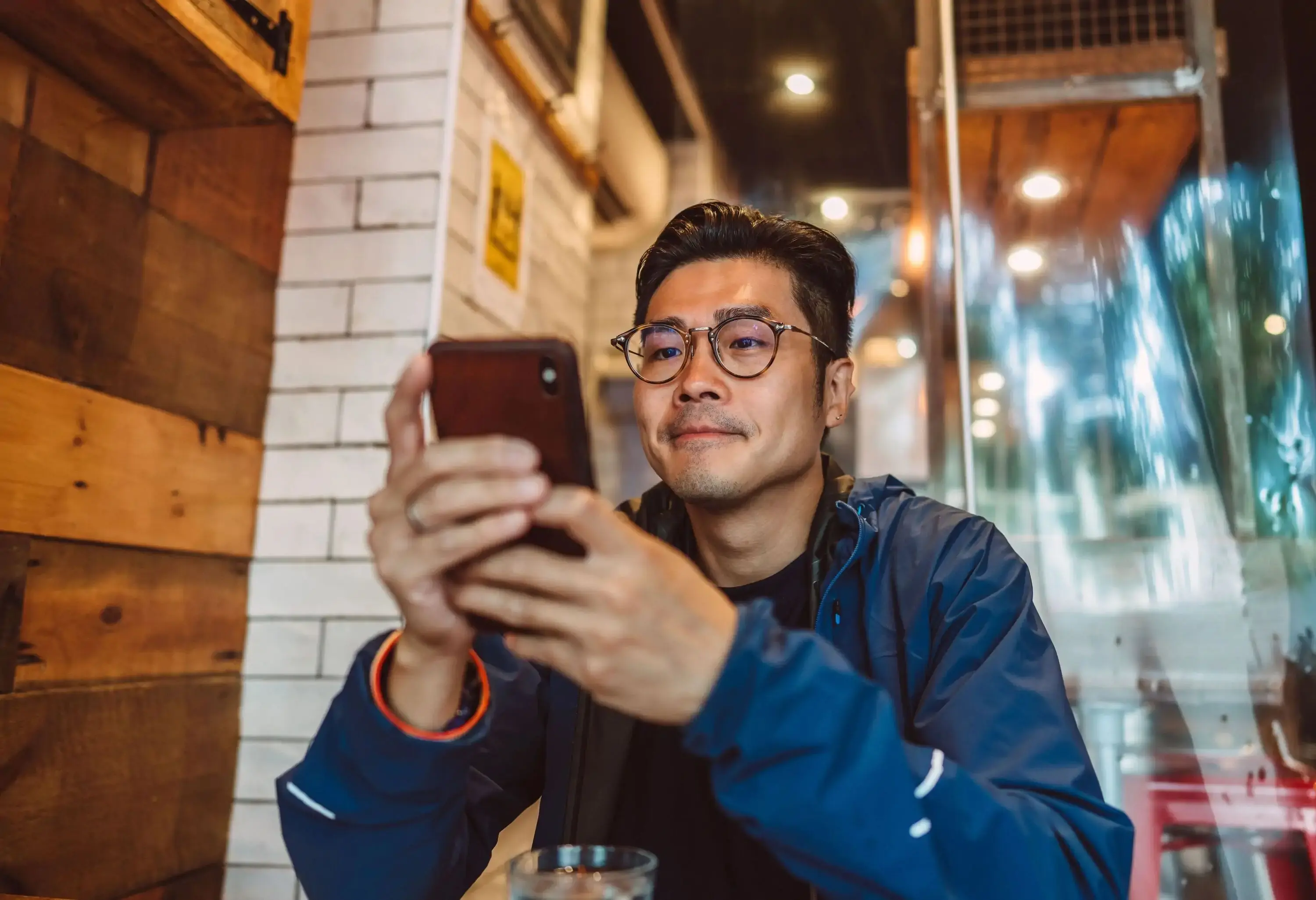 A focused man with round glasses, wearing a blue jacket, holds a smartphone in both hands and looks at its screen.