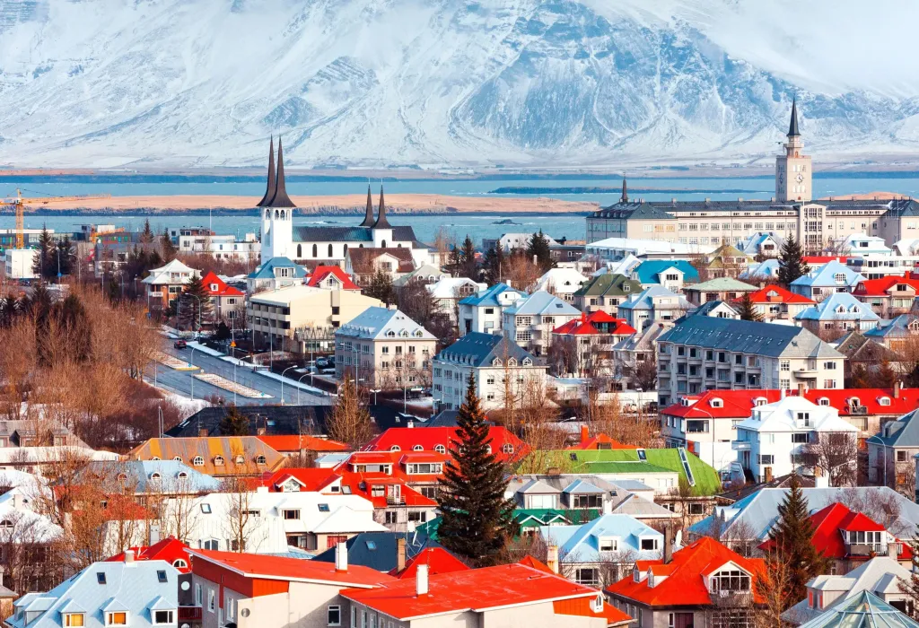 Building spires dominate a colourful cityscape along a blue bay with an ice-covered steep mountain in the background.