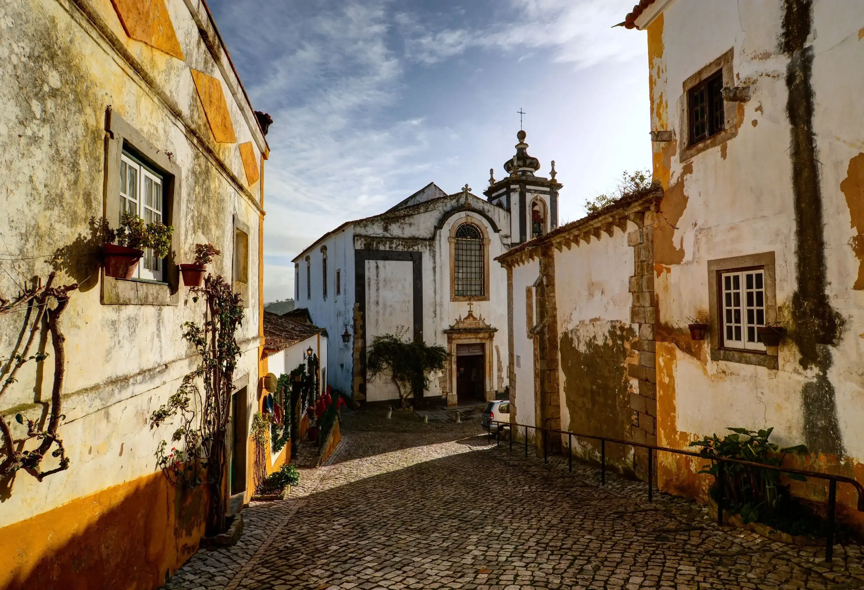 A narrow cobblestone street along old buildings towards a white church with a bell tower.