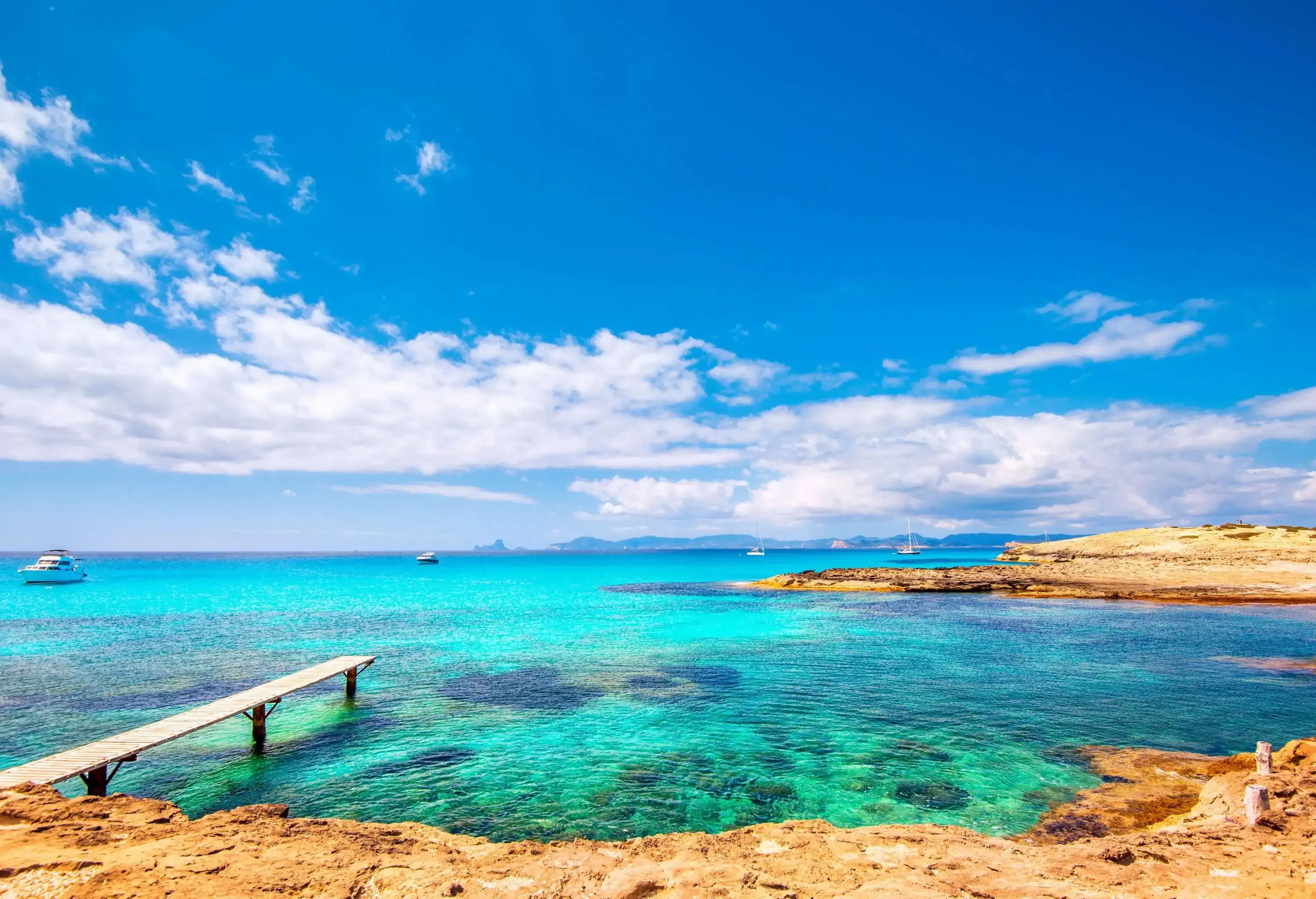 A wooden jetty on a pristine beach with yachts floating in the ocean.