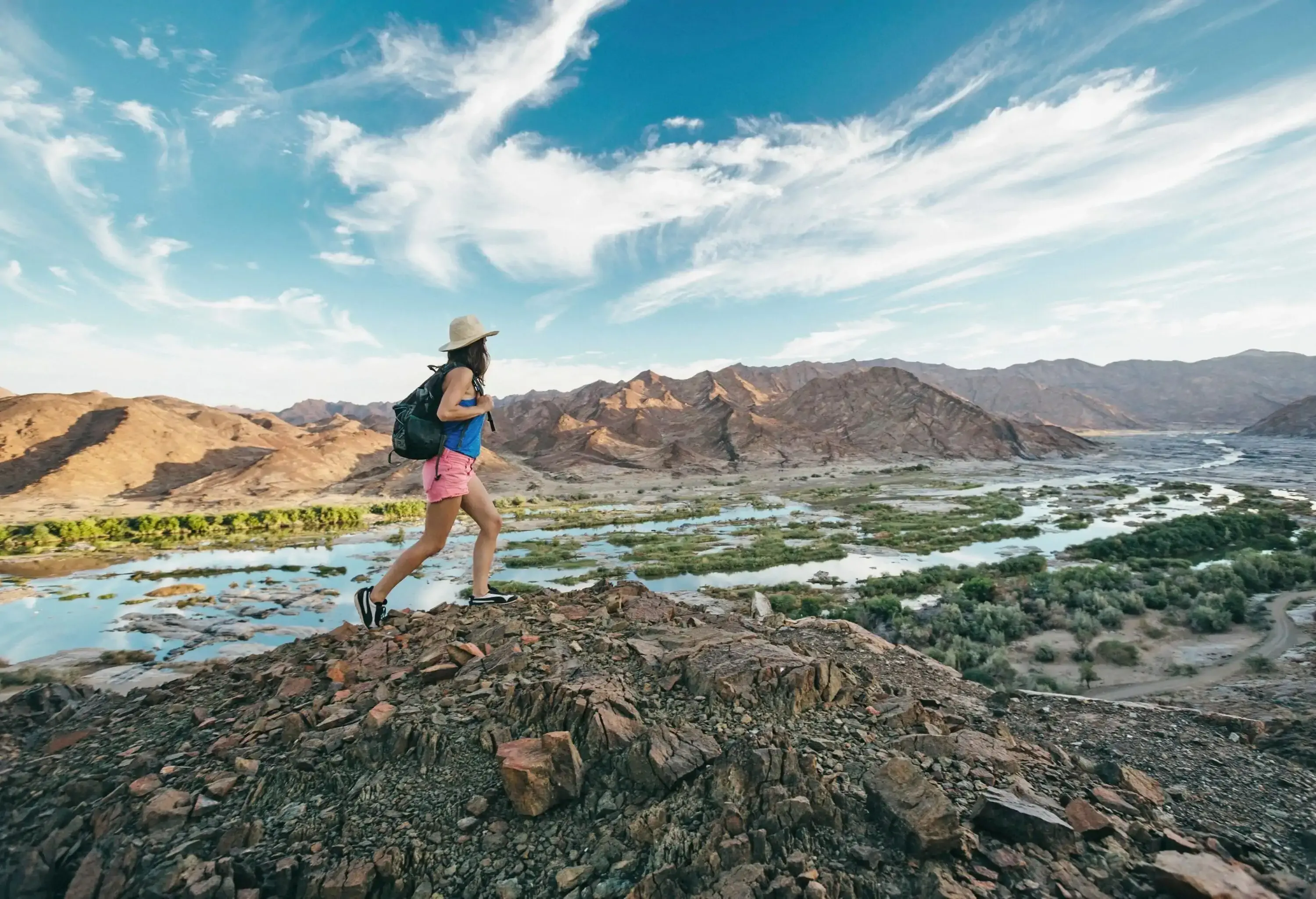 A woman with a hat and bag traverses a rock ridge while gazing at the river along the mountains.