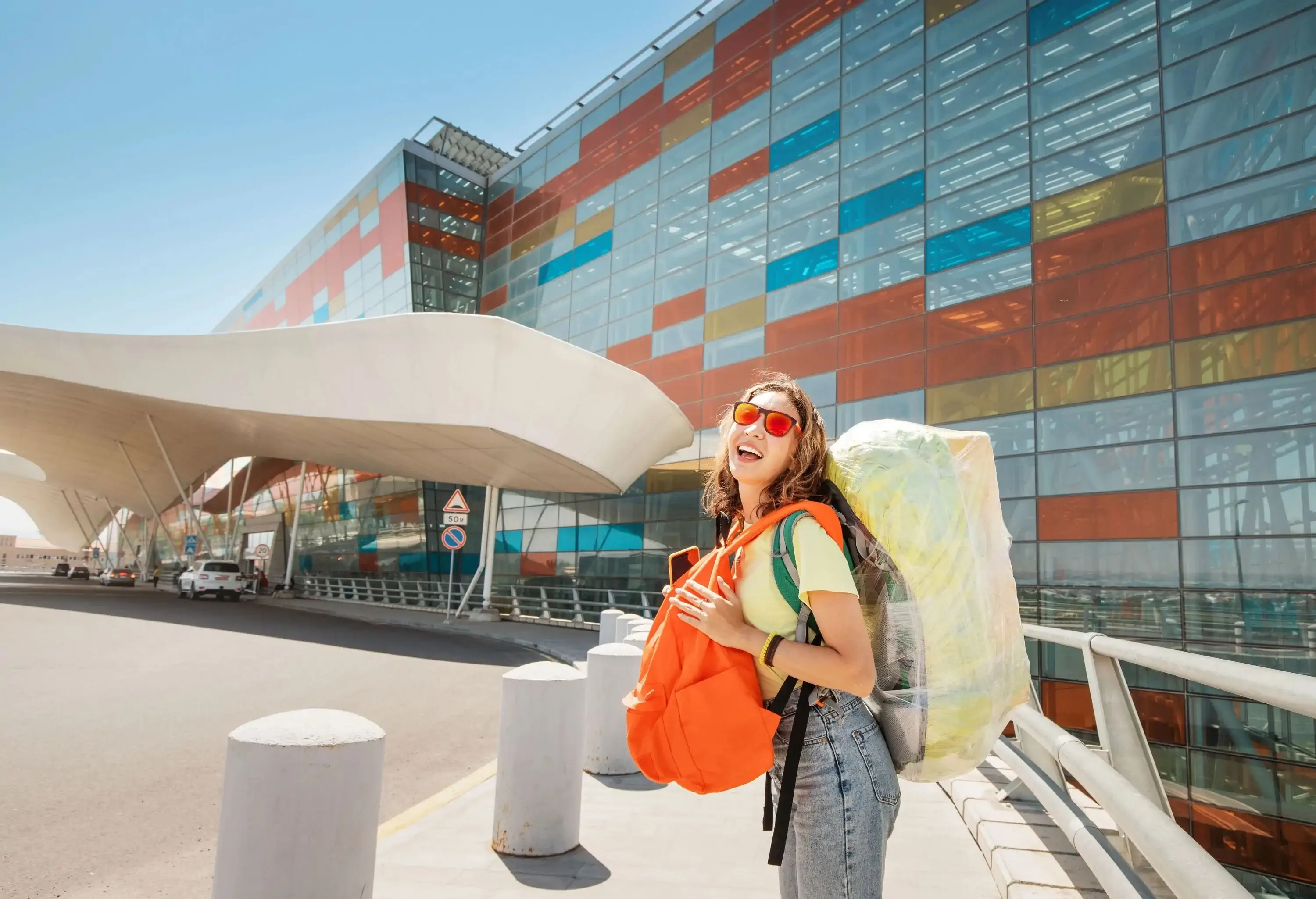 A smiling traveler with two large backpacks stands outside a modern building with a colorful facade under a blue sky.