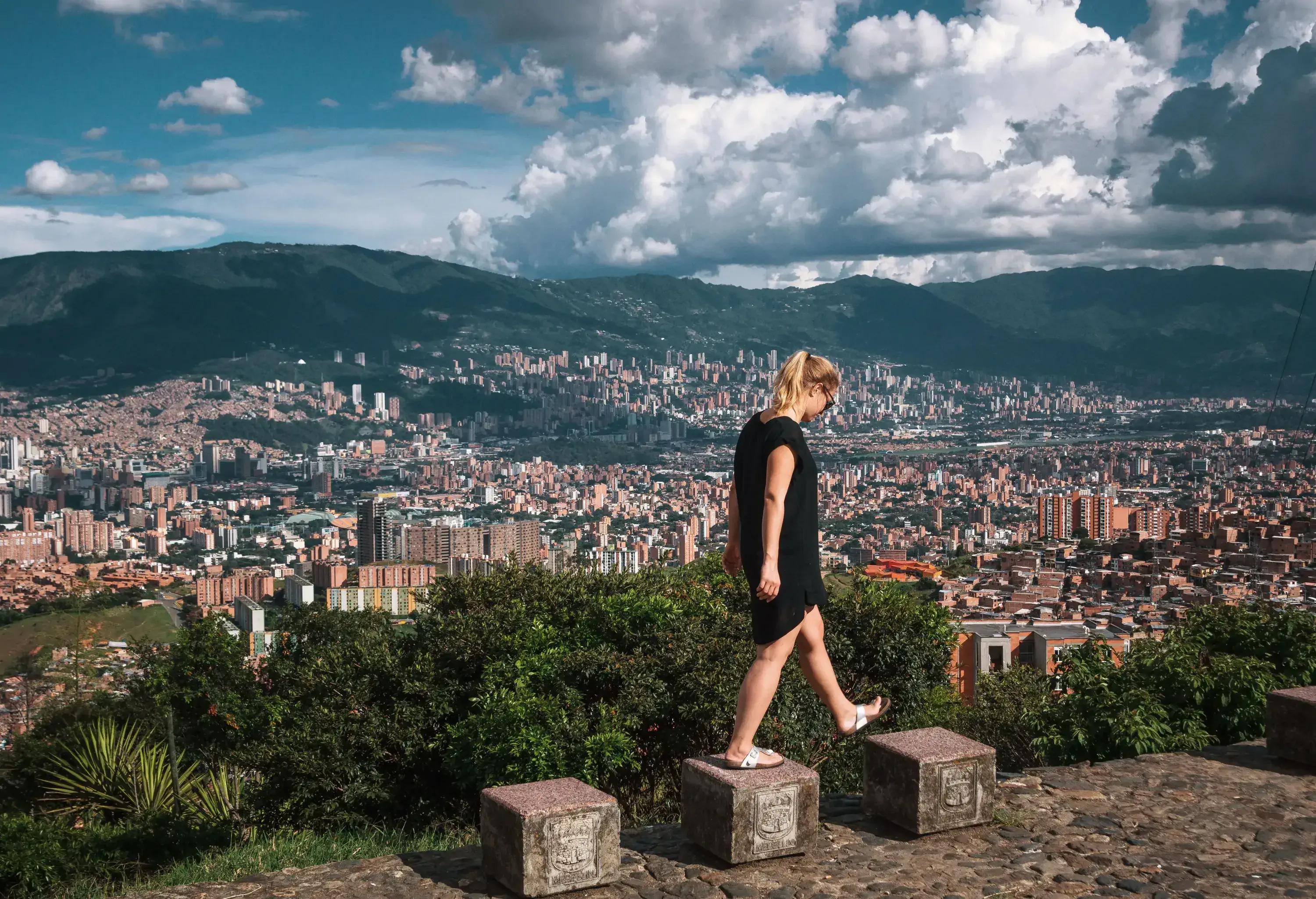 A person in a black dress is joyfully skipping on blocks, overlooking a vast cityscape below and a majestic mountain range in the distance.