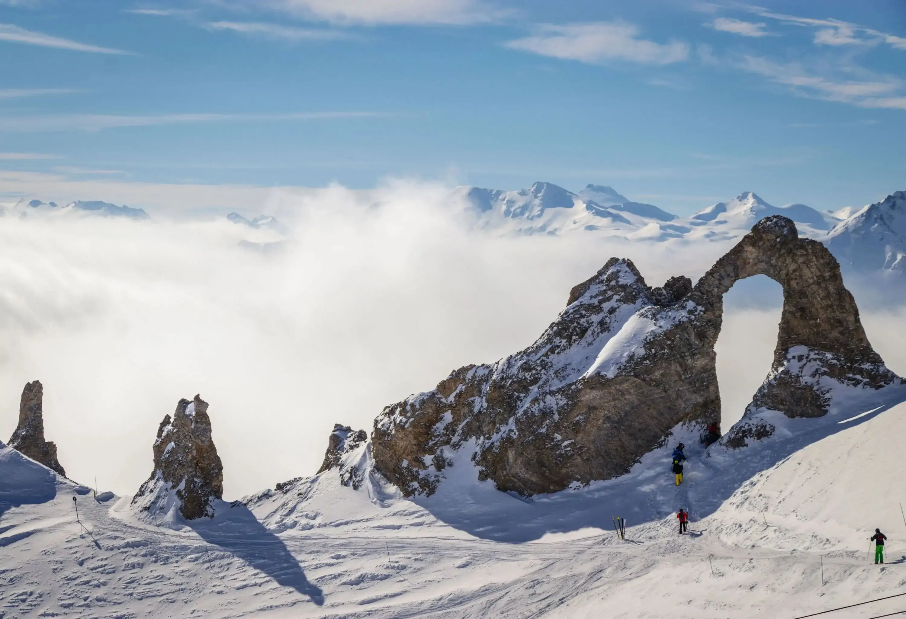 People ski towards a large hole of an arched rock formation with views of nearby valleys covered in fog.