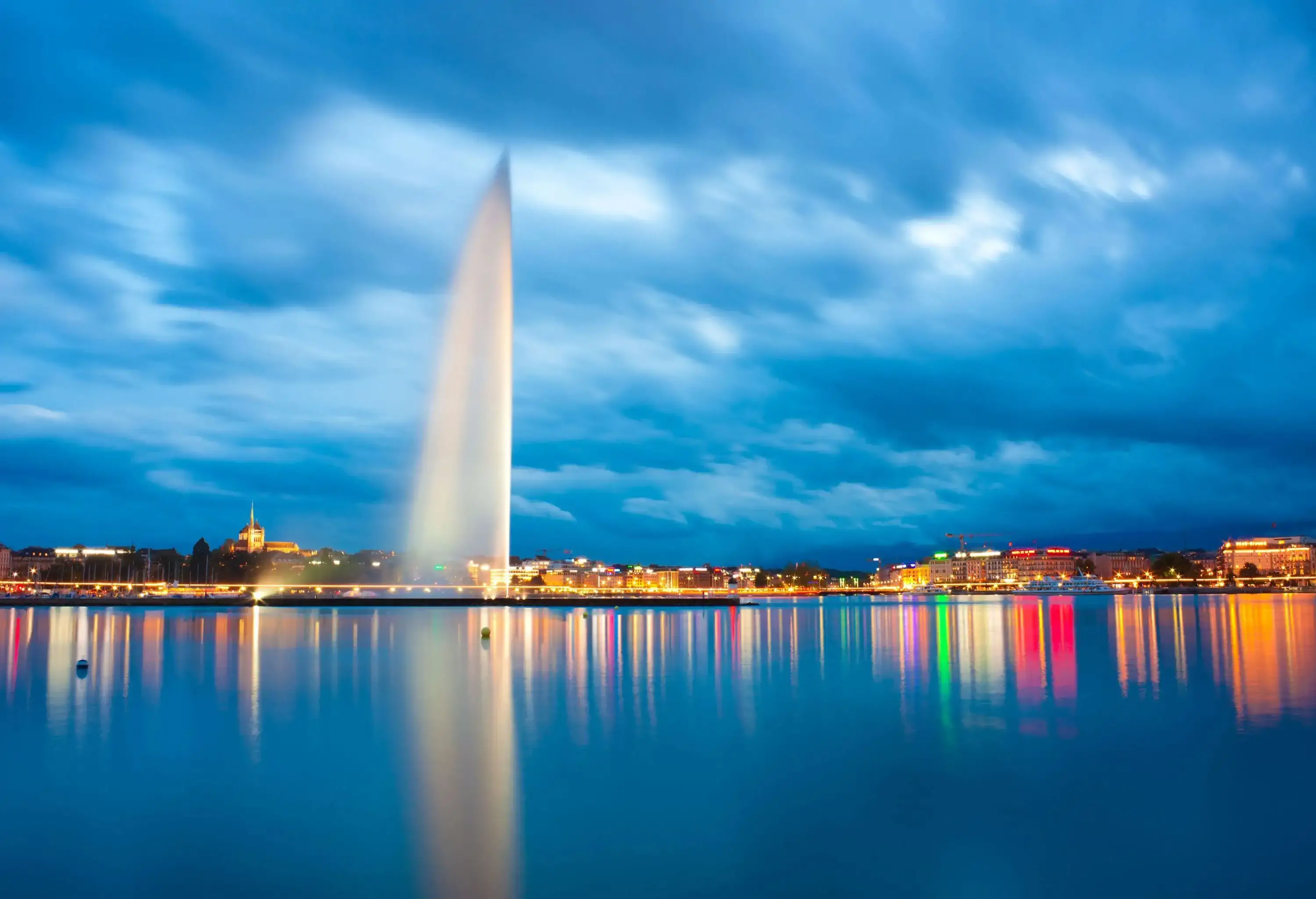The Jet d'Eau jutting into the sky as streaks of night lights reflect on the lake.
