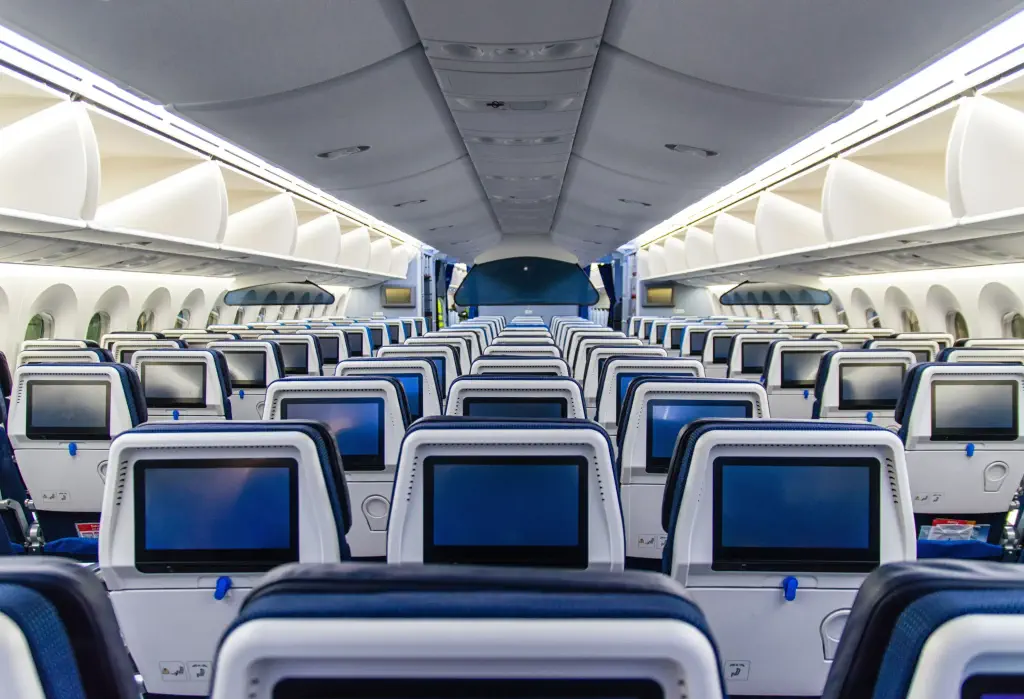 Rows of airplane seats with touchscreen TVs in a cabin.