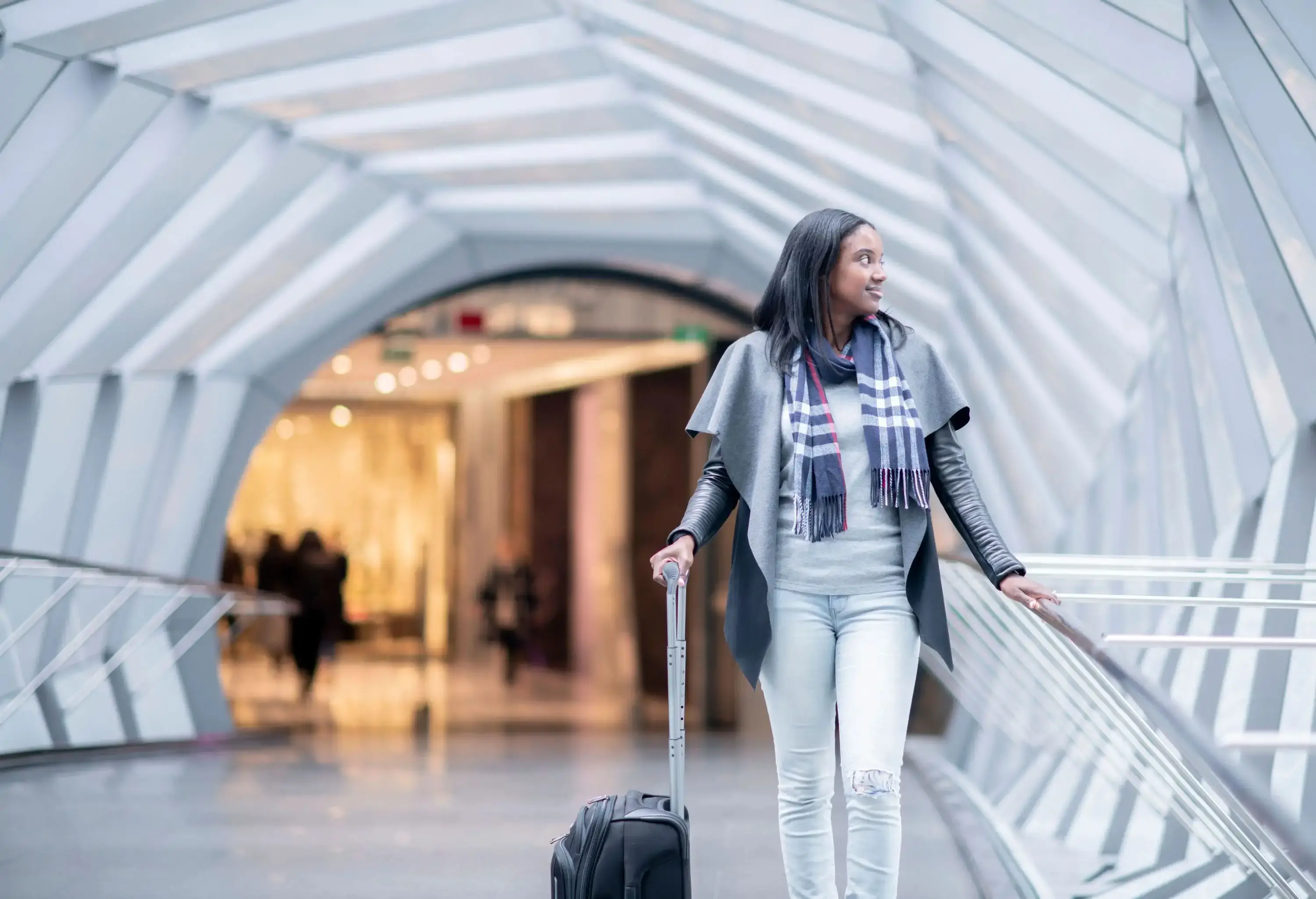 A woman wearing a scarf walking through a modern walkway with geometric designs while pulling a suitcase.