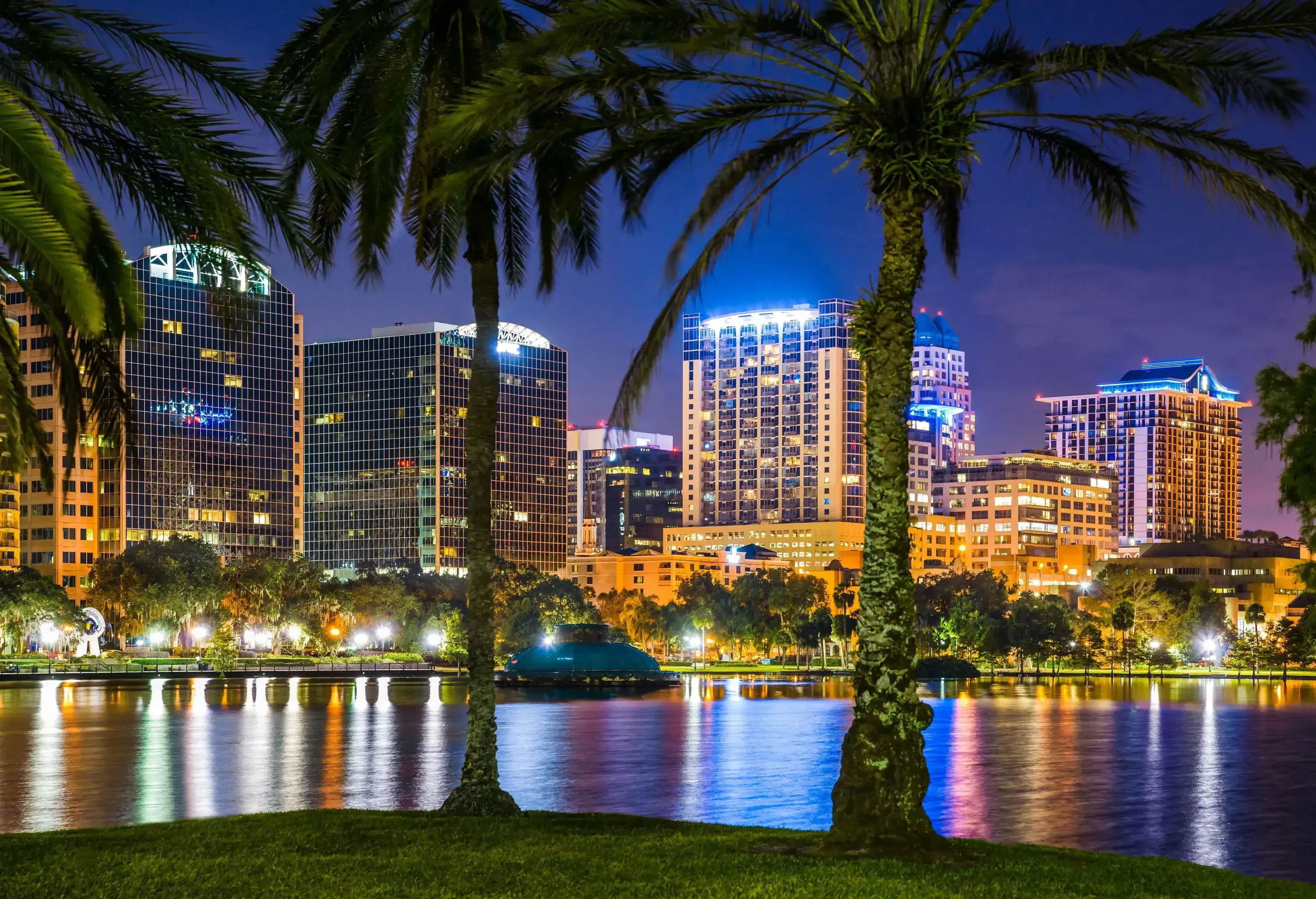 A lake at night with a dome-shaped fountain surrounded by lights, trees, and skyscrapers.