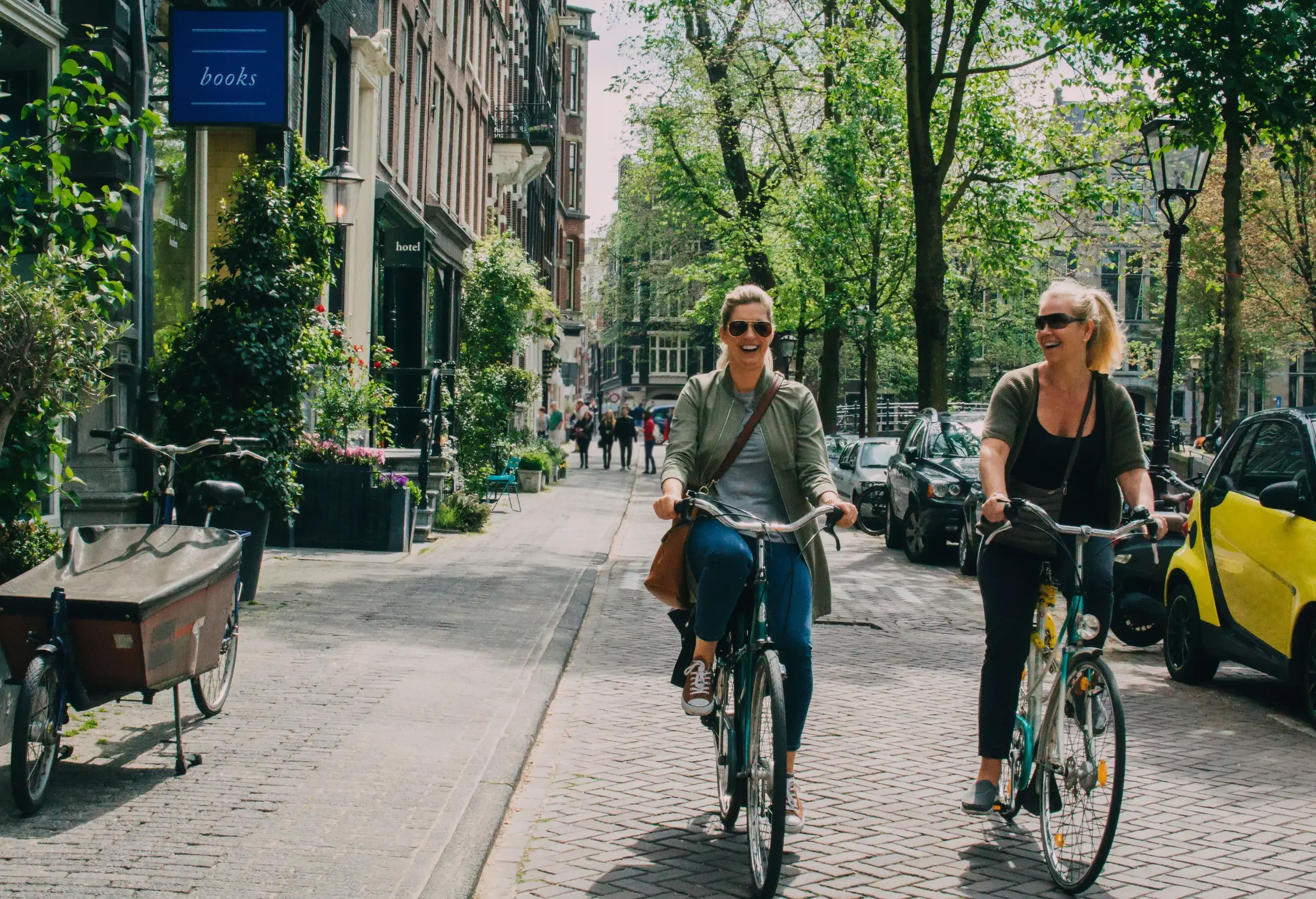 Two women riding their bikes on a pavement lined with trees and cars.