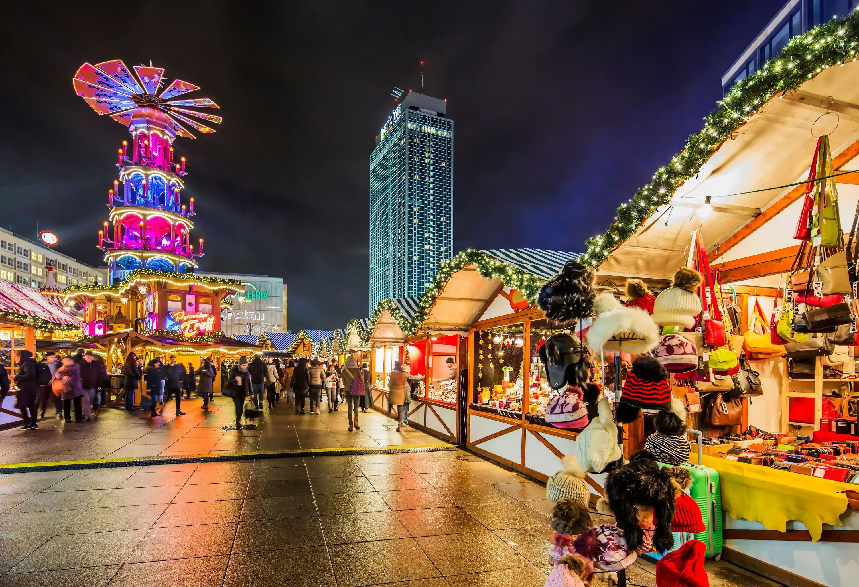 Night view of a bustling Christmas market with brightly lit stalls and a tall illuminated tower in the background.
