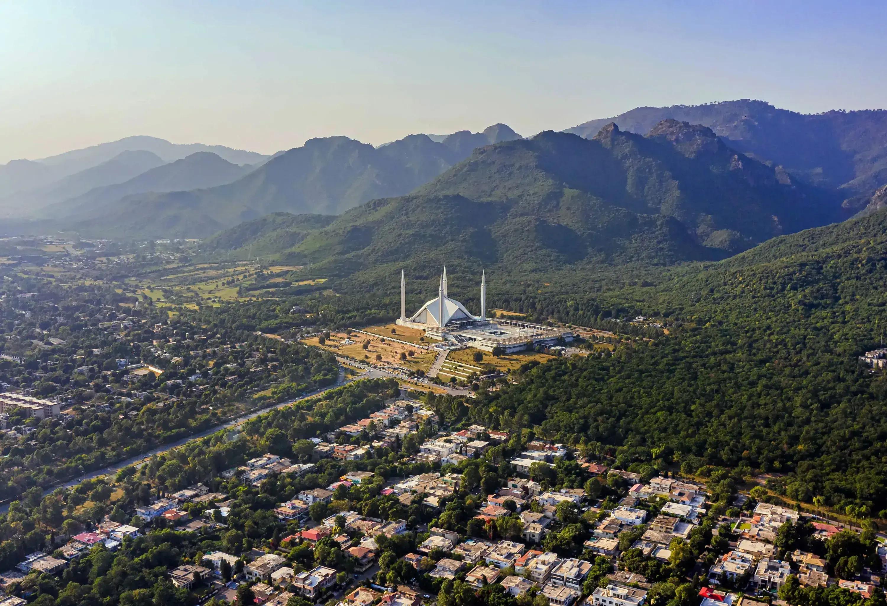 A white mosque with four minarets at the base of a forested hill next to a residential area amongst lush trees.