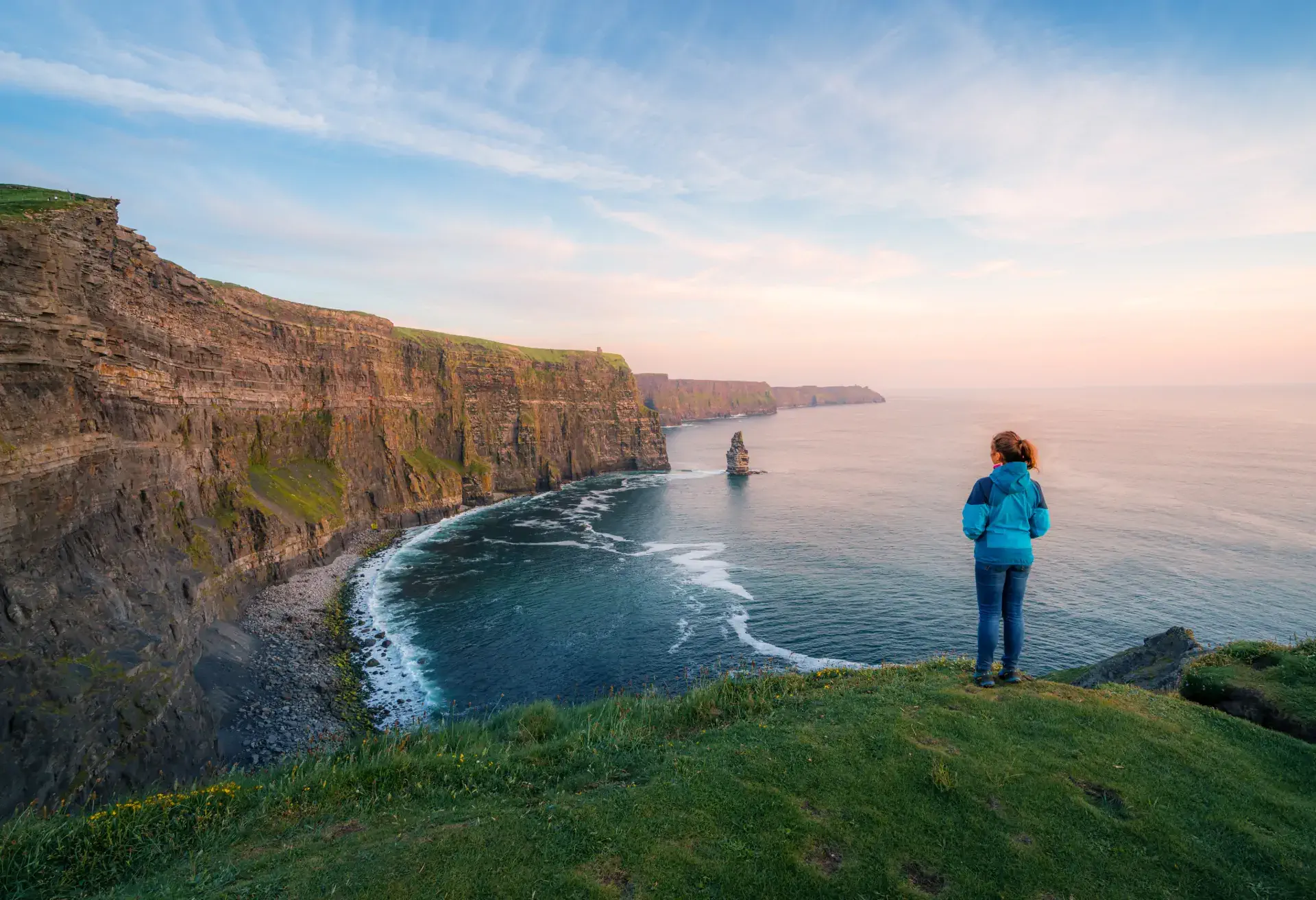 Person standing on cliff edge overlooking ocean and tall sea cliffs at sunset.