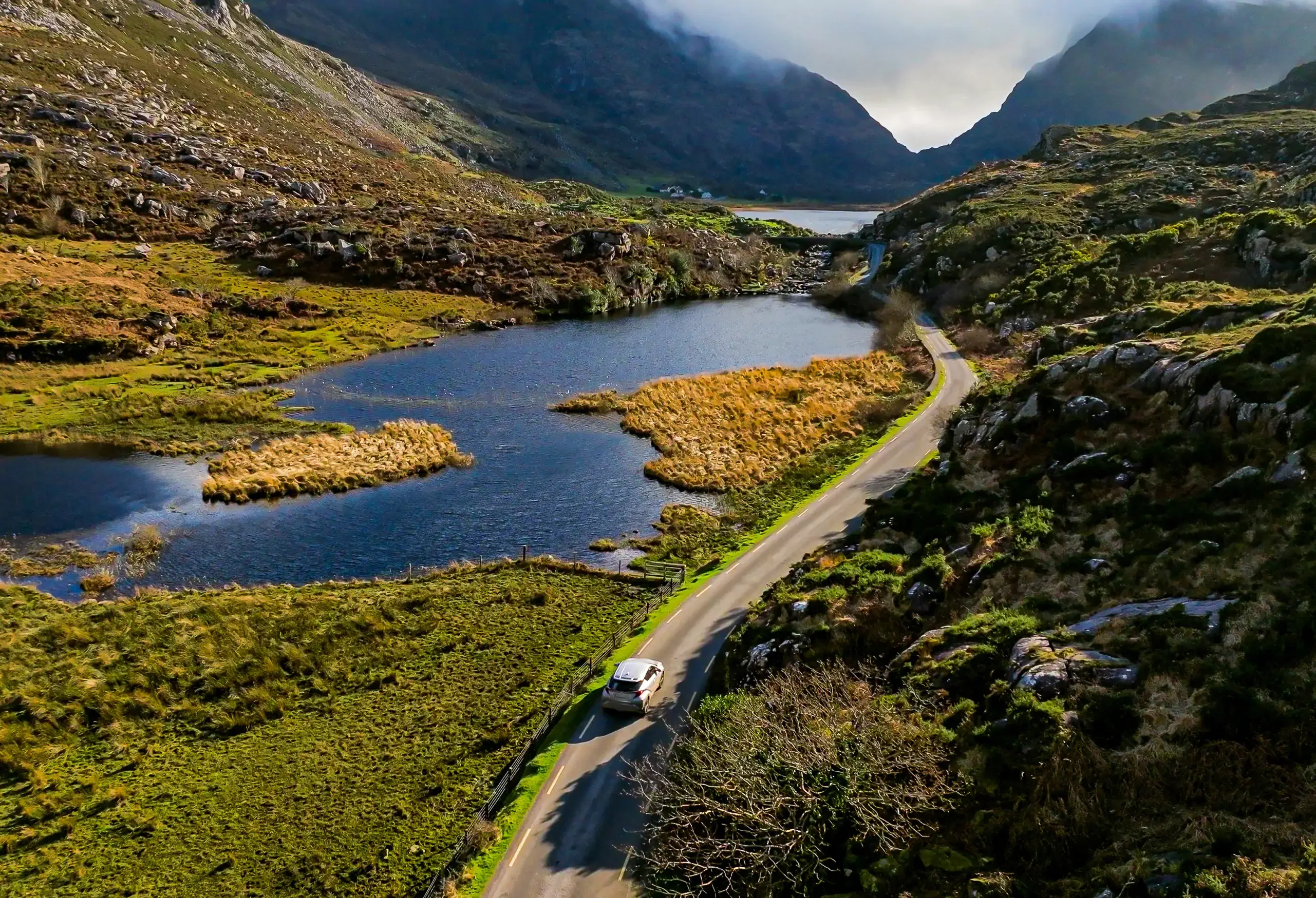 Car driving along a winding road beside a lake surrounded by green mountains.