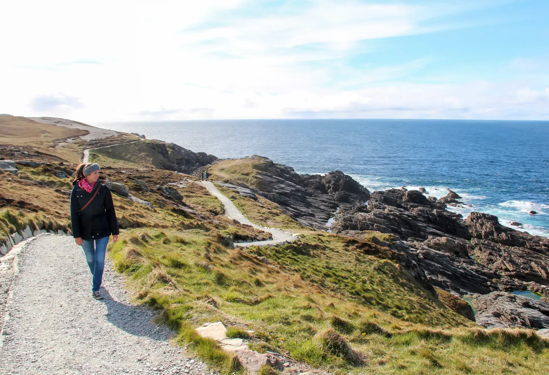 Person walking on a coastal path with cliffs and ocean under a bright blue sky.