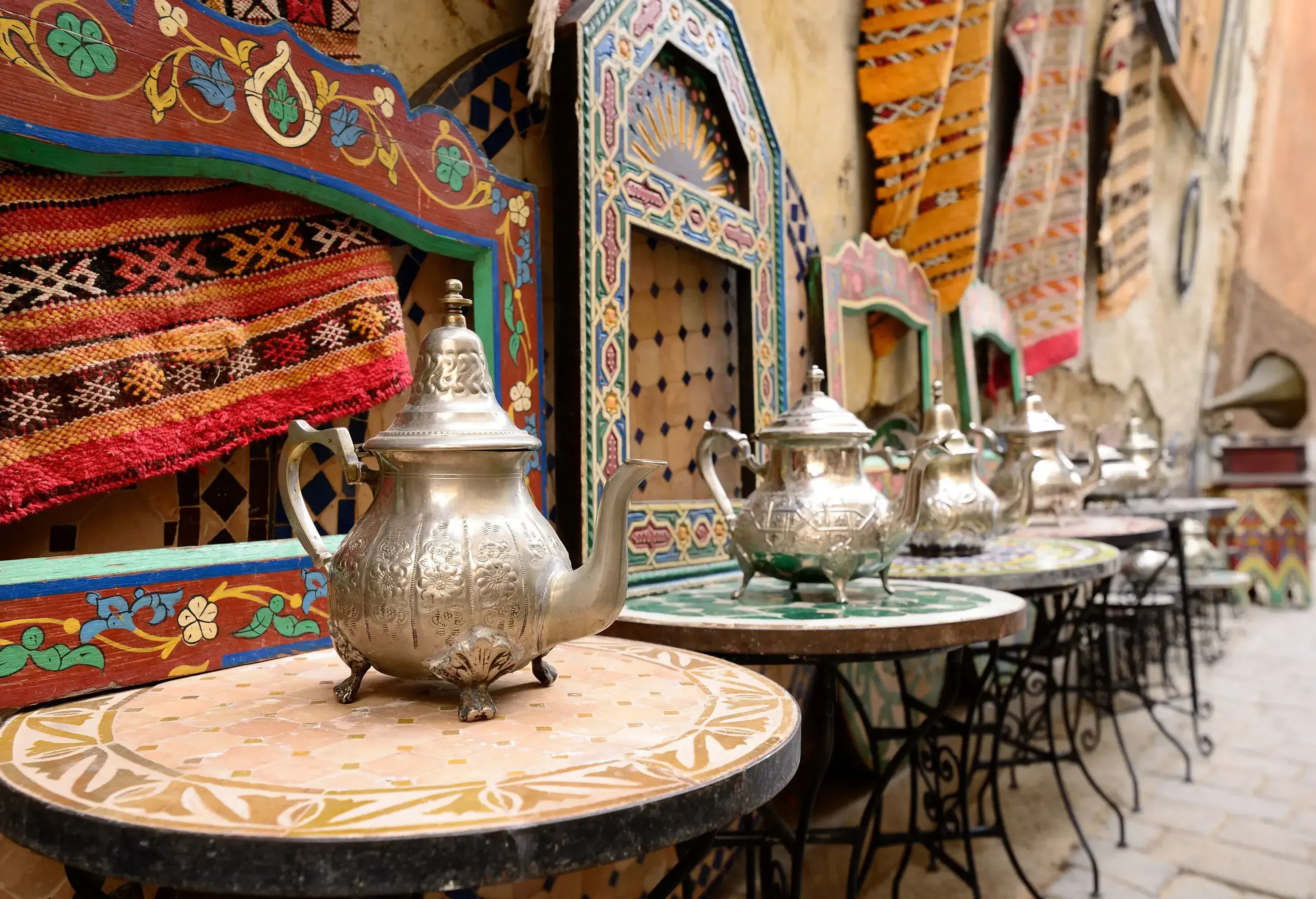 A row of tables with stainless steel kettles along a display of colourful carpets.