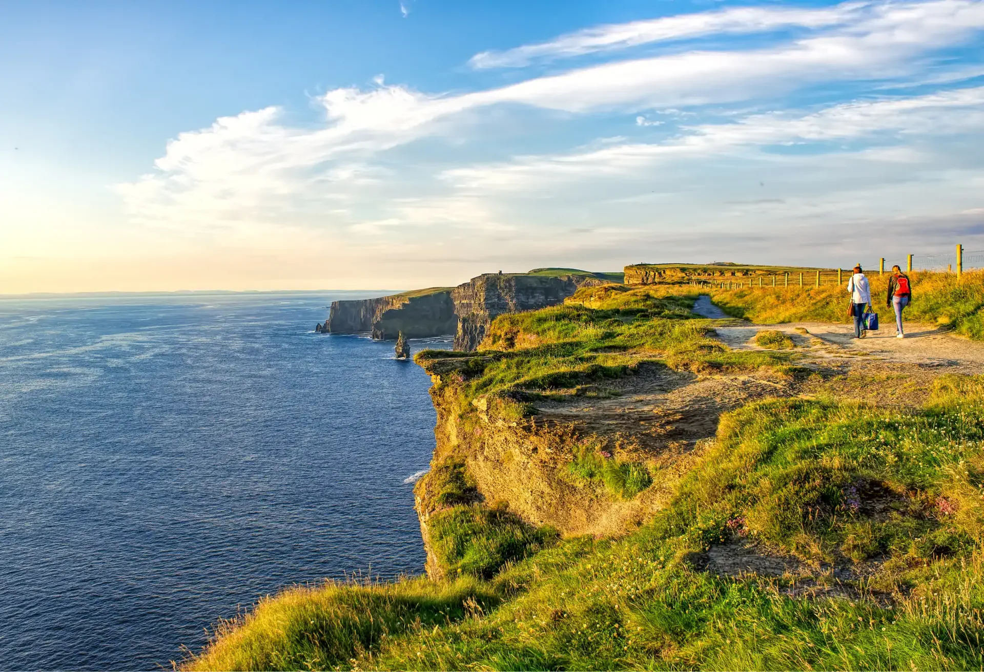 People walking along grassy cliffs overlooking the ocean at sunset