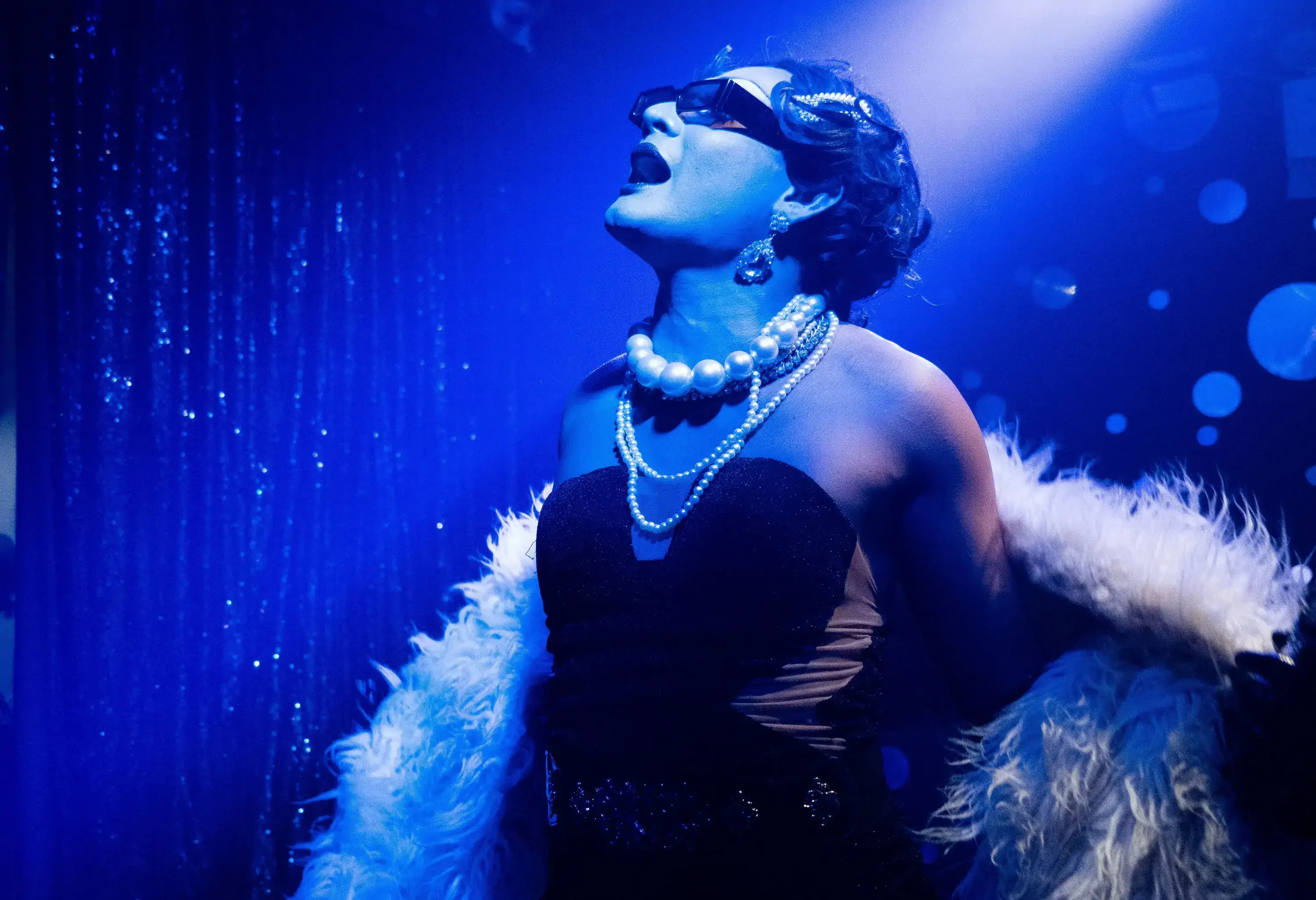 A drag performer in black dress, feathered cape, and pearl necklaces sings on a stage with blue lights.