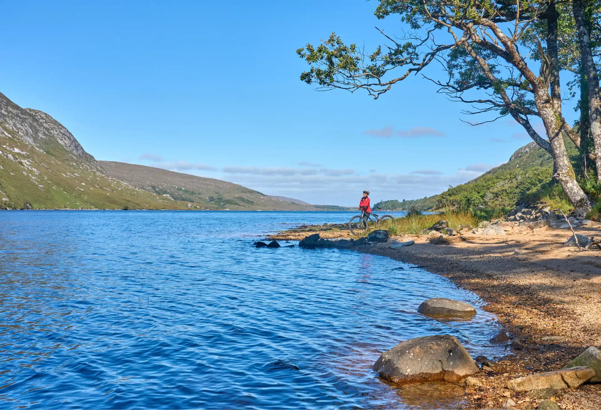 Cyclist resting by a calm lake with hills and trees under a clear blue sky.