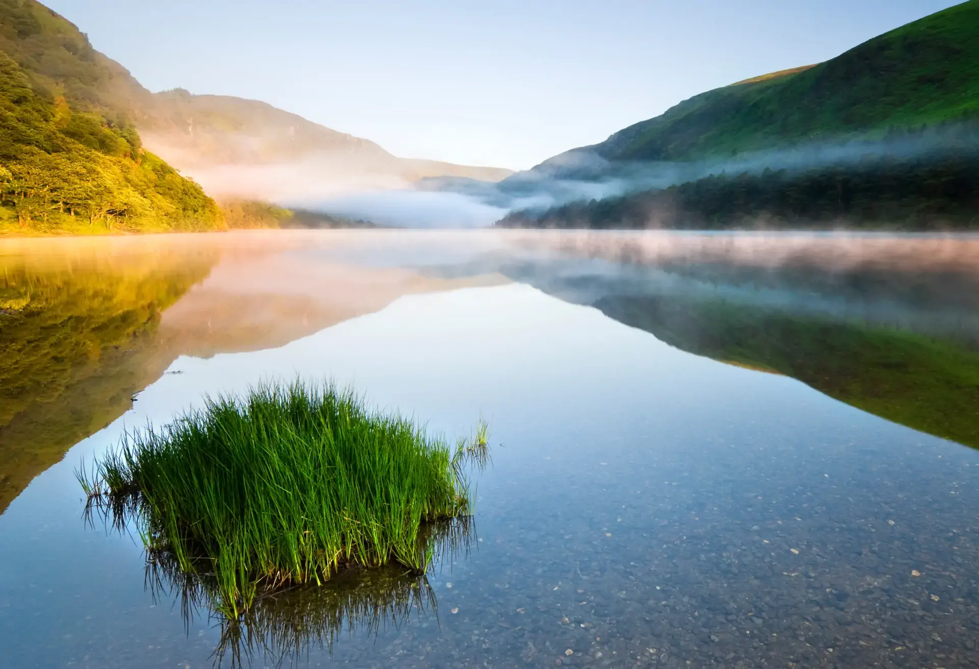 Calm lake reflecting misty hills and green grass at sunrise in a serene valley.