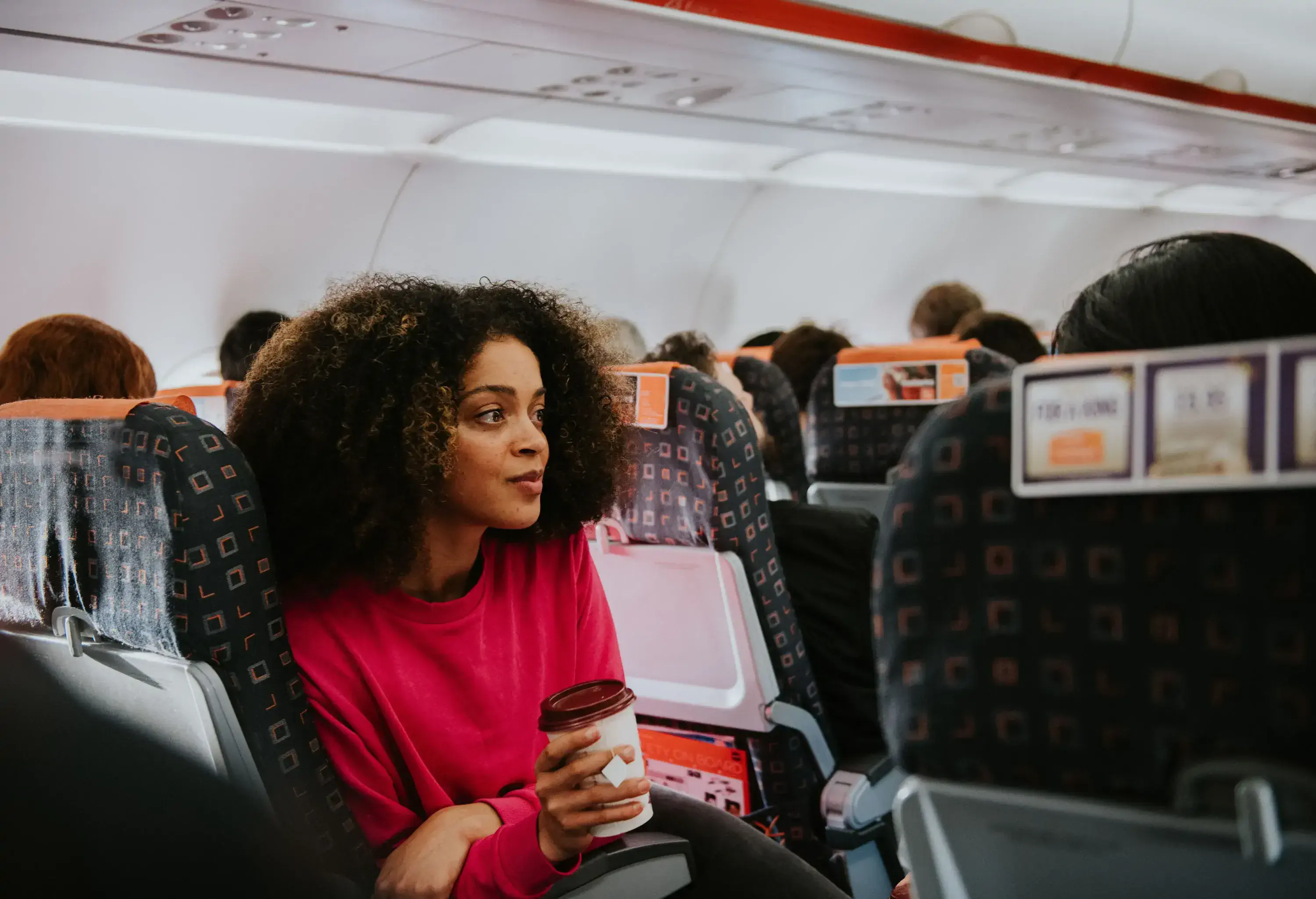 A woman in a pink sweater, sitting on an airplane while holding a coffee cup.