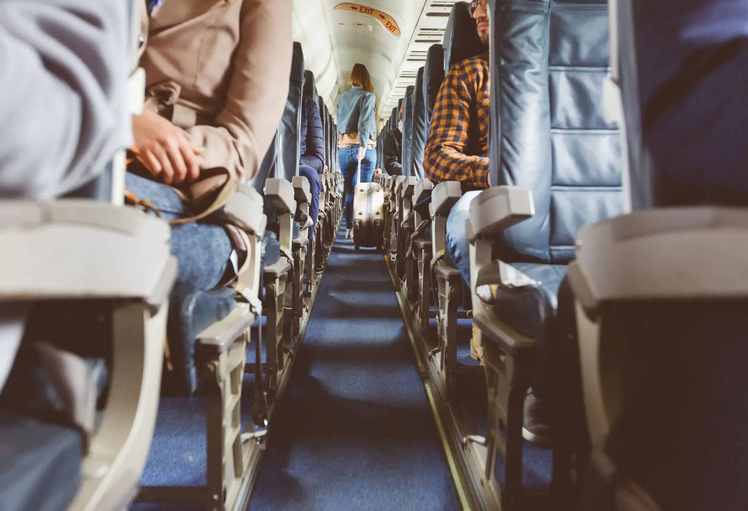 A long blonde woman walks on the aisle and pulls her luggage to her seat in an airplane.