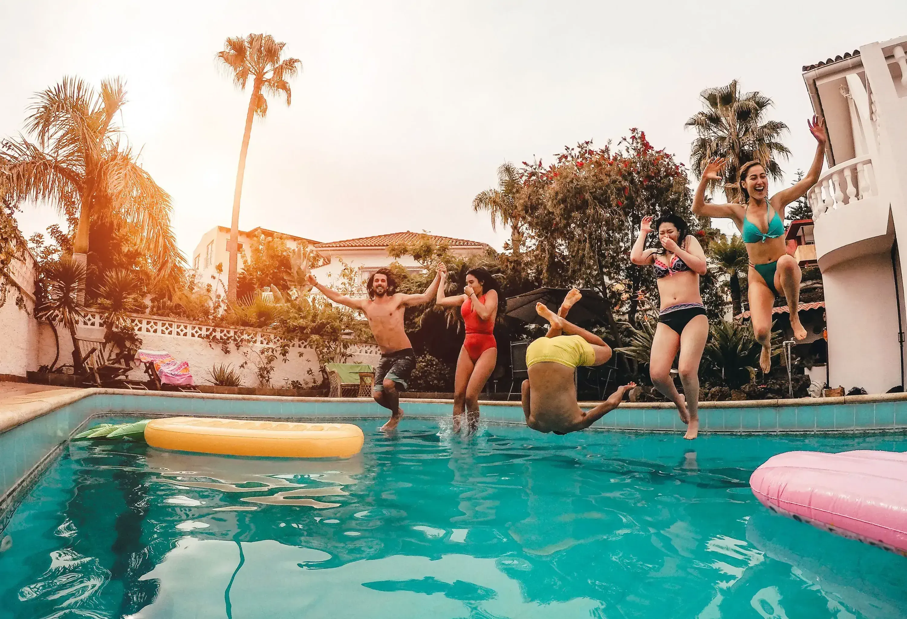 A group of friends in swimwear jump into a swimming pool of a tropical house.