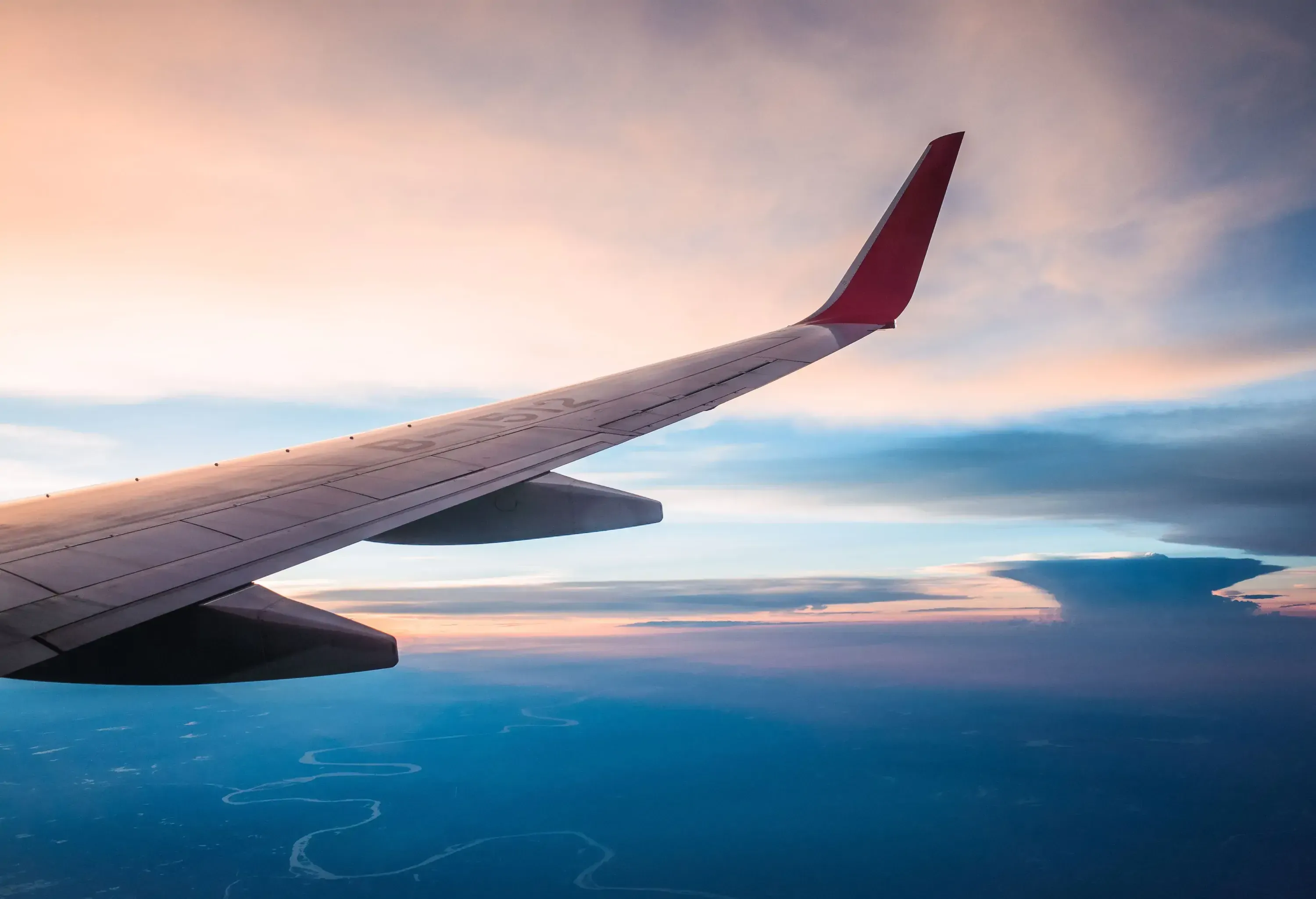 Through the plane's window, a breathtaking pink and blue cloudscape unfolds, with the wing of the aircraft adding a sense of perspective to the awe-inspiring aerial vista.