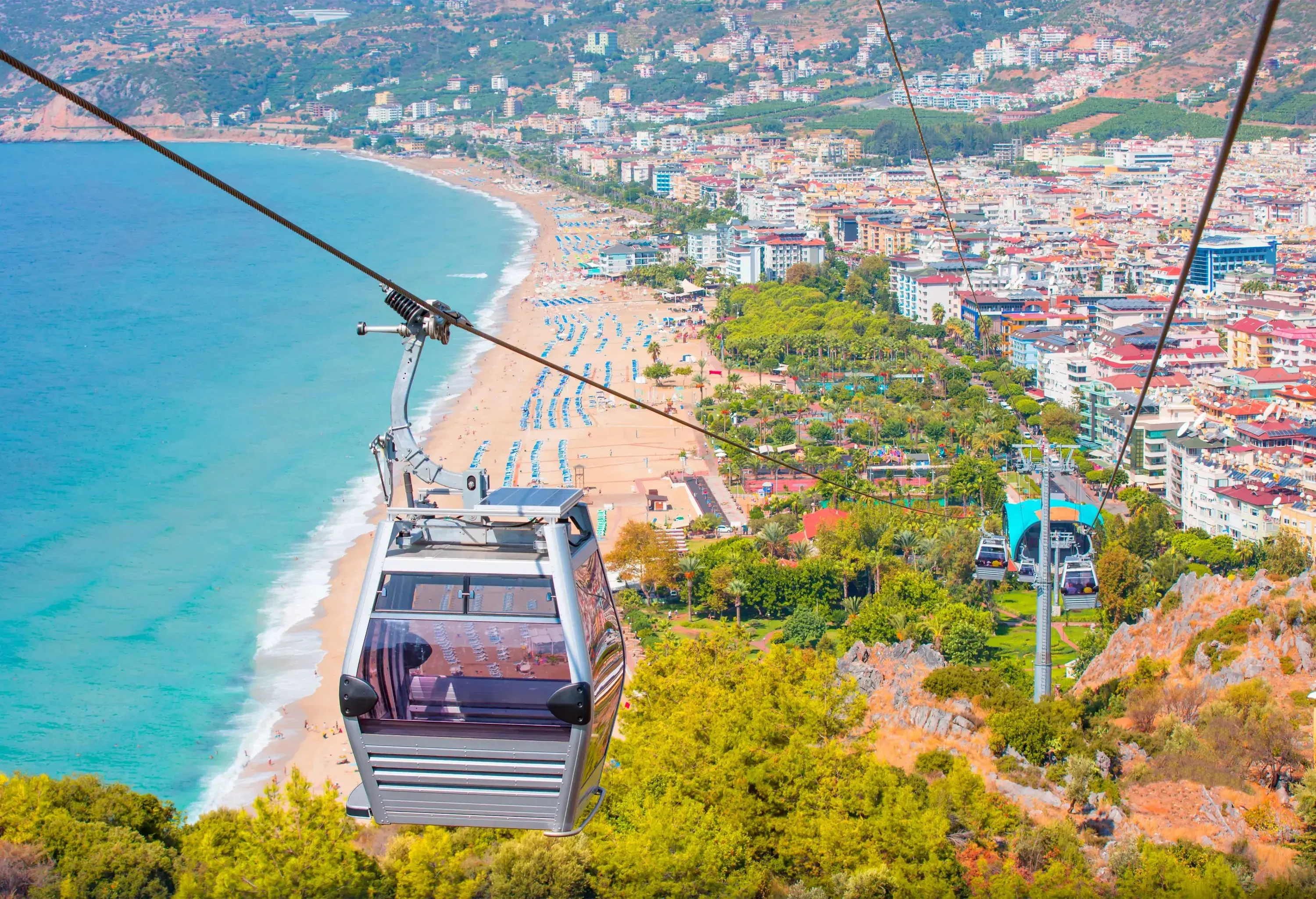 Cable cars ascending and descending a lush, wooded slope, while a bustling beach and city below offer a contrasting urban energy to the serene natural beauty of the surroundings.