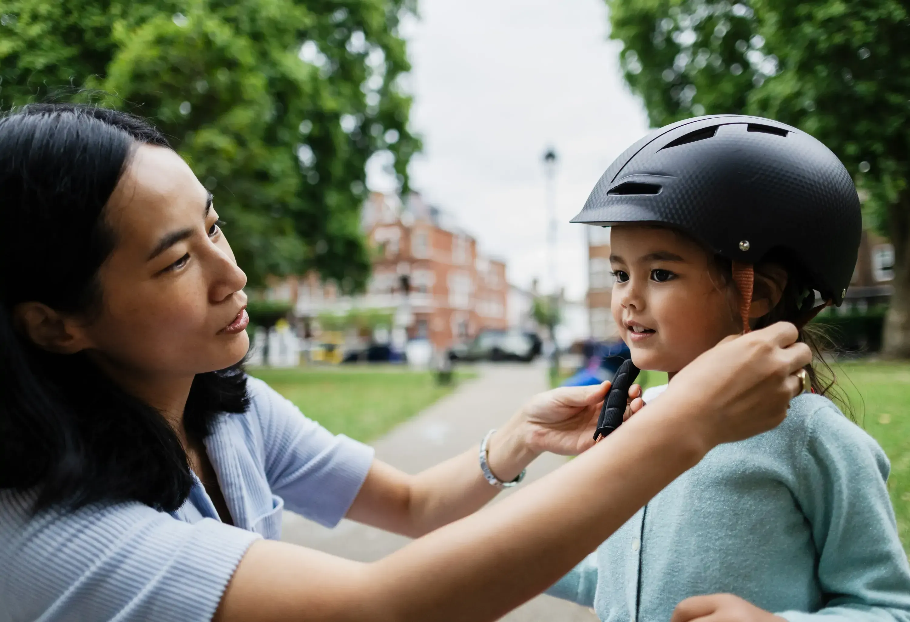 Asian mother secures her daughter's bike helmet, ensuring her safety before cycling in a park