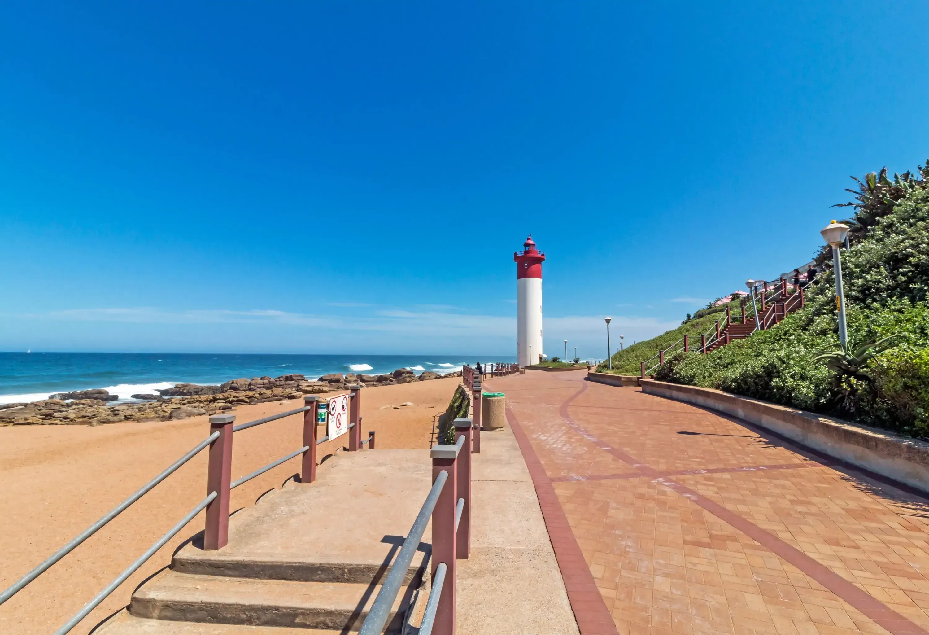 Concrete stairs on a beachfront promenade that leads to a red and white lighthouse overlooking a vast ocean.