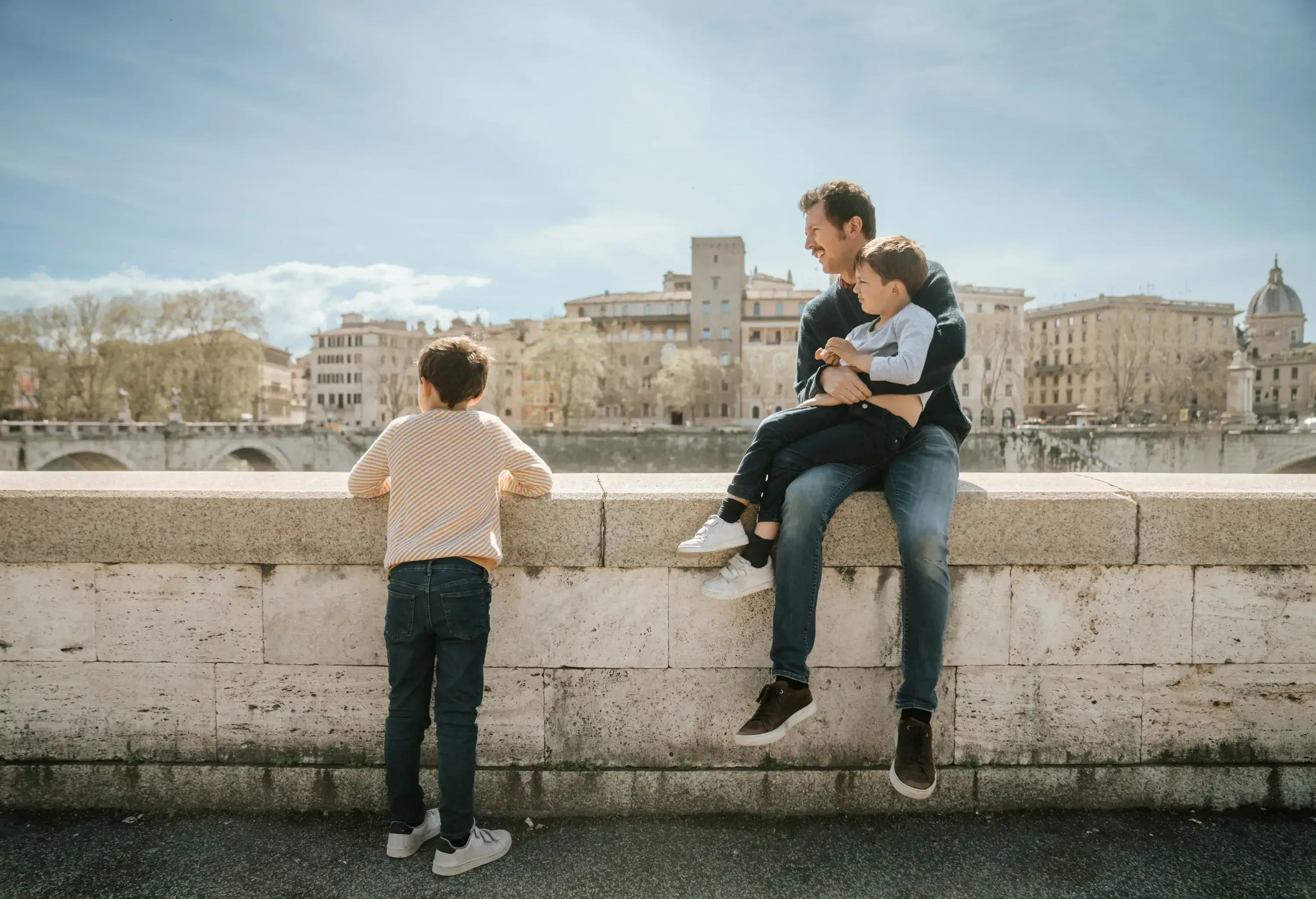 Father and two sons enjoy a sunny day on a riverbank. One child leans on the stone wall while the father, seated, holds the younger son, smiling against an urban backdrop.