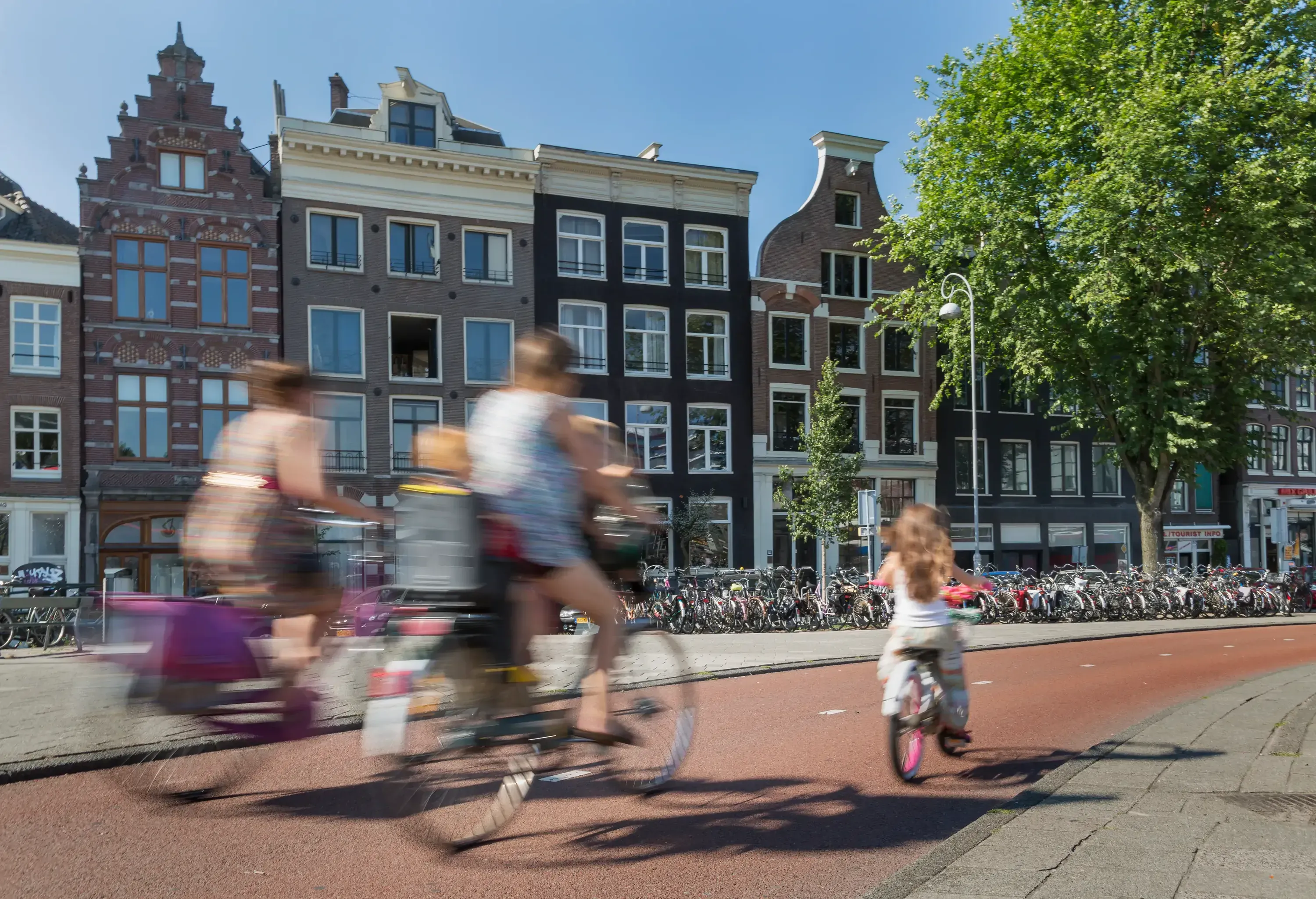 Blurred cyclists ride past traditional Dutch canal houses in Amsterdam. A child follows on a bike, conveying a lively, dynamic urban scene.