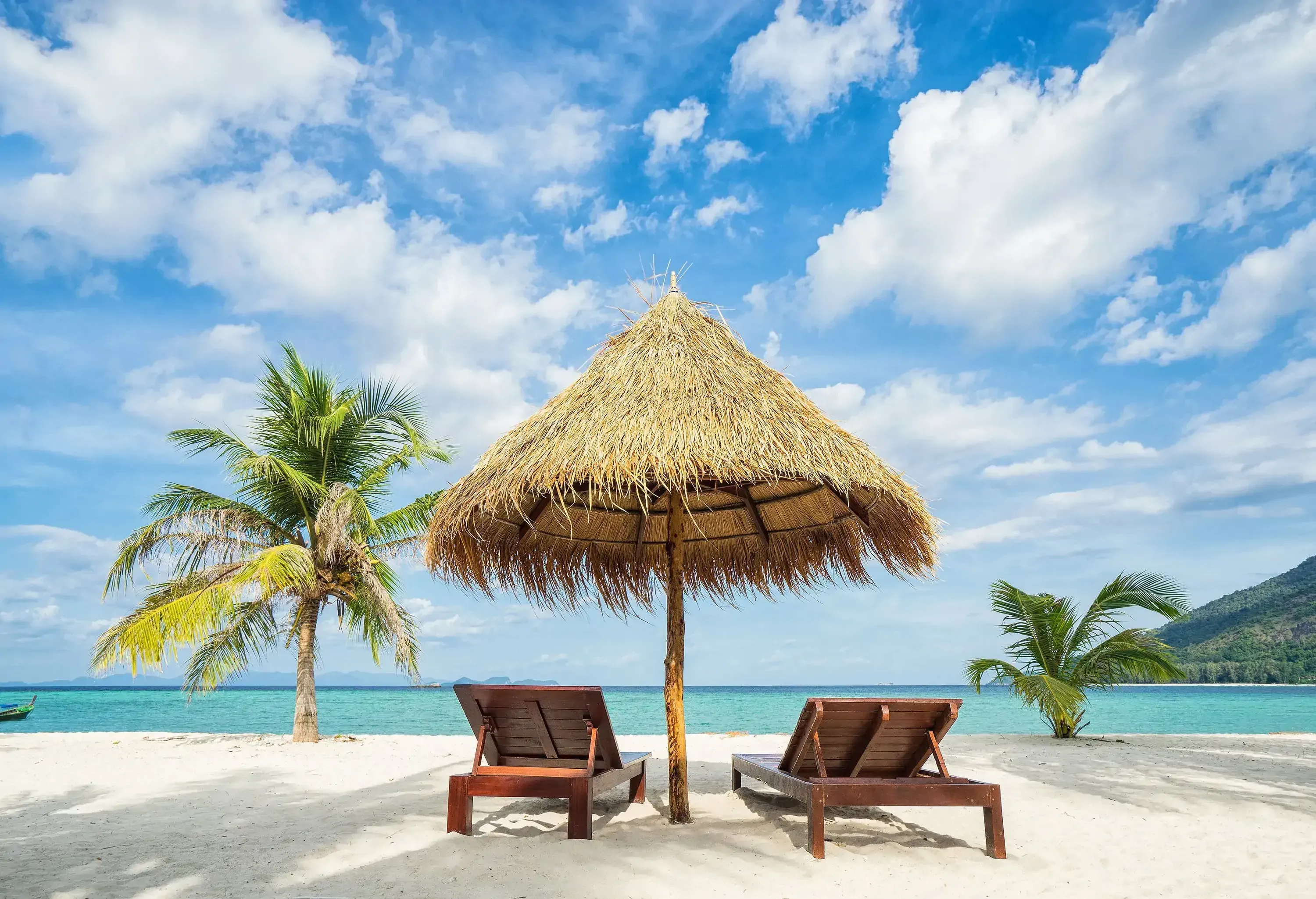 Two wooden sun loungers under the shade of a nipa hut on the shore of the turquoise sea under the cloudy blue sky.