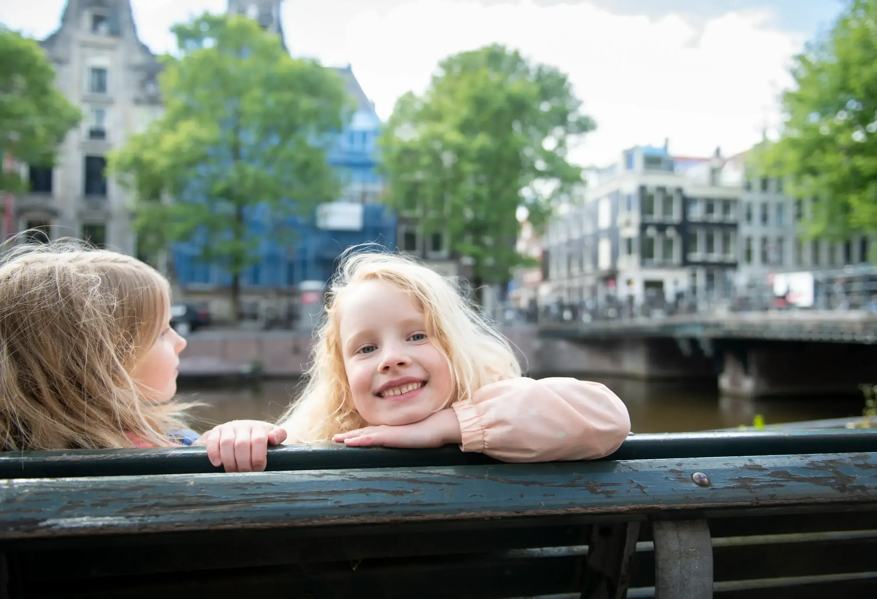 Two young girls with long blonde hair lean over a green railing by a canal in a city, with historic buildings and leafy trees in the background.