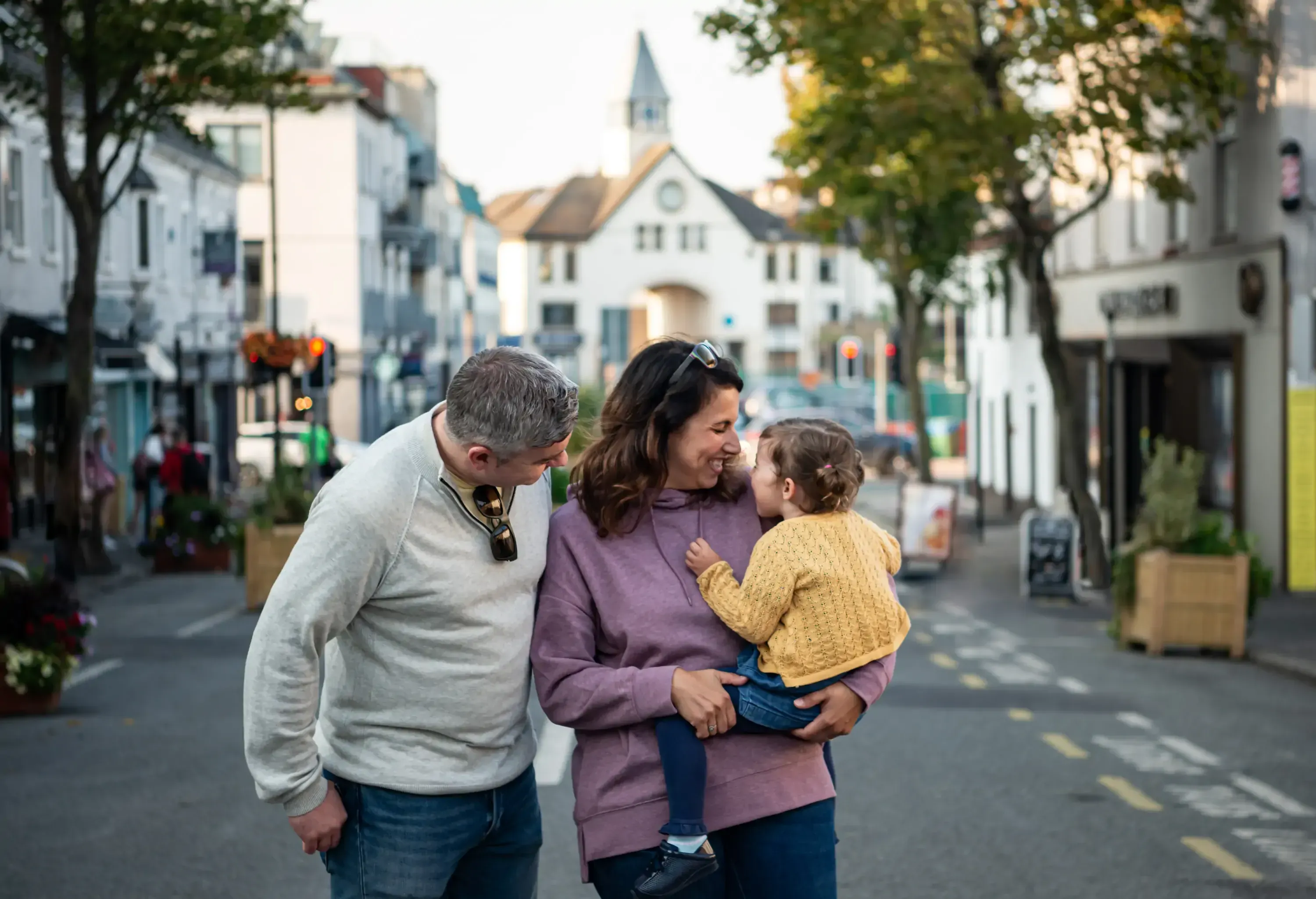 A family of three shares a joyful moment on a quaint street. A woman holds a toddler, smiling at each other, while a man stands beside her. Sunlit street and historic buildings in the background.