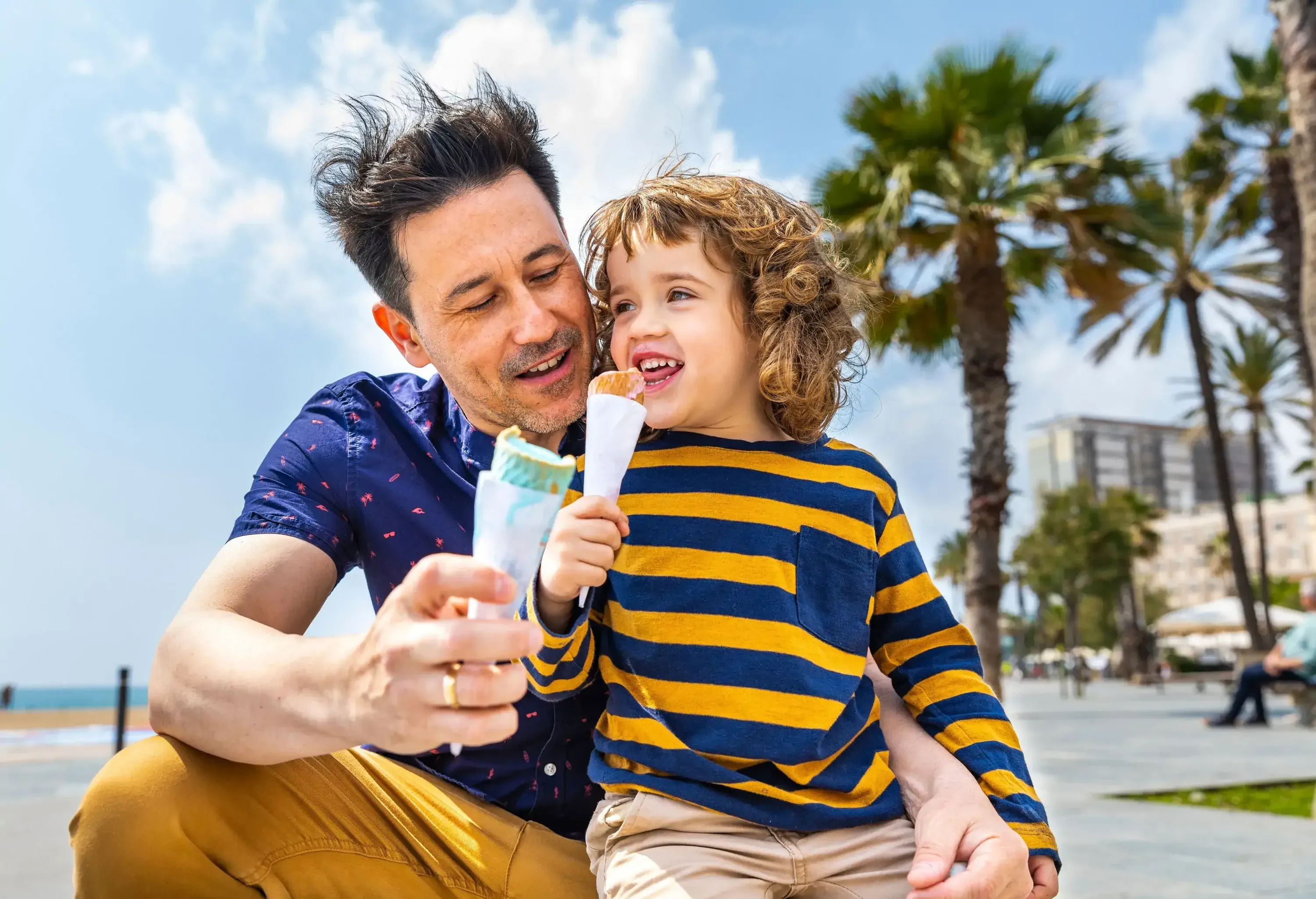 A man and a child joyfully hold ice cream cones outdoors near palm trees under a bright sky.