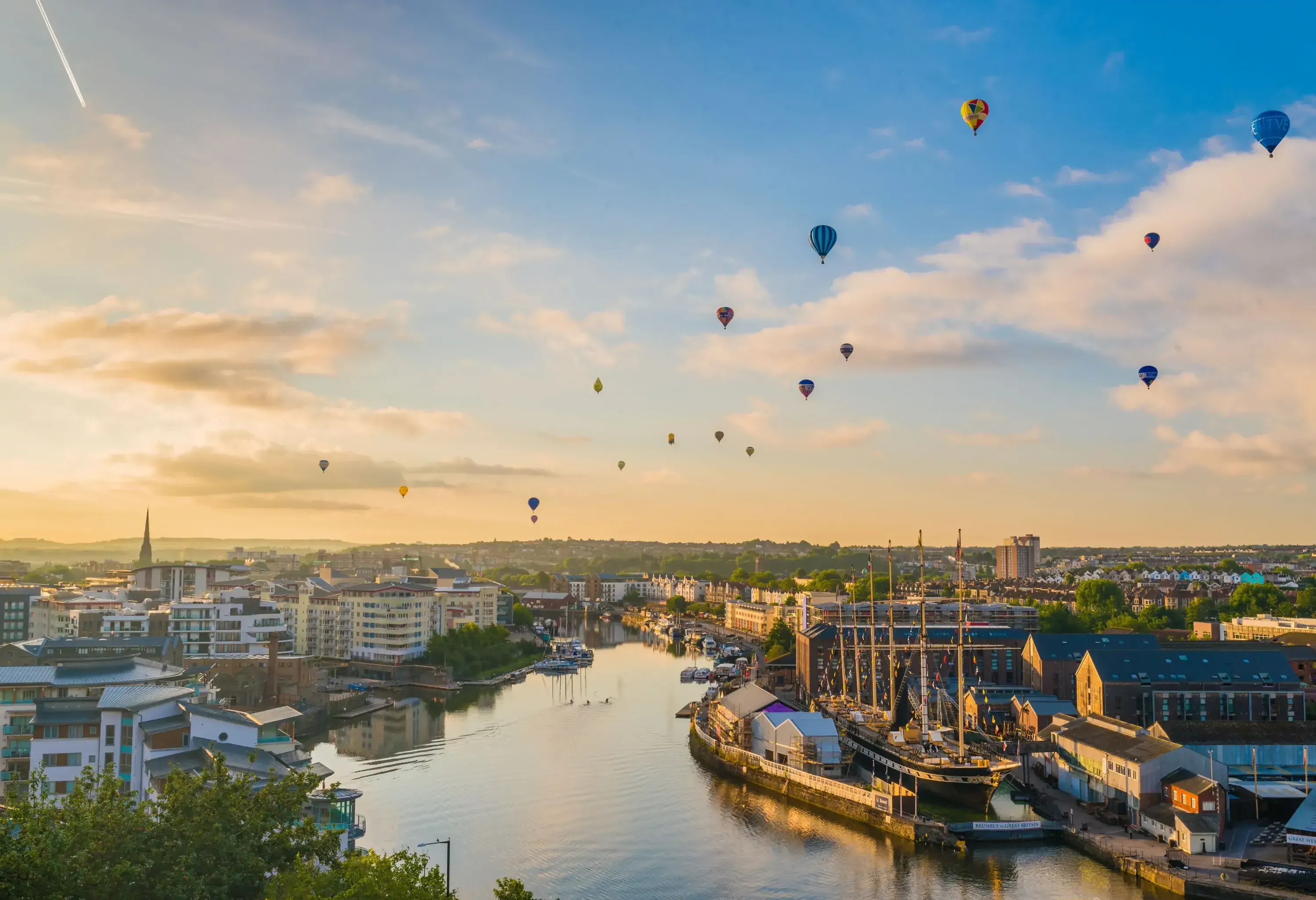 Hot air balloons flying above the buildings along the banks of a river.