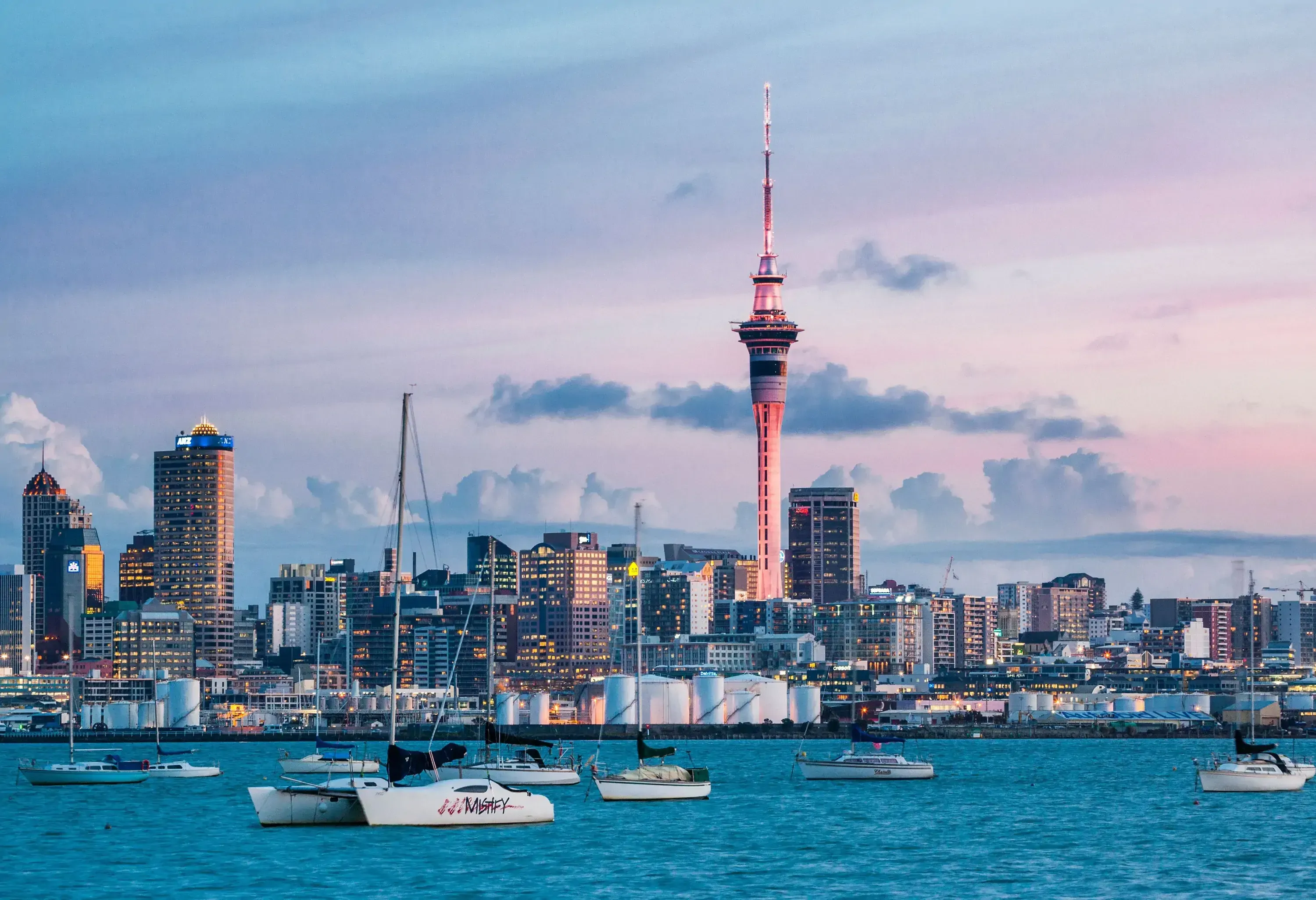 A brightly lit coastal urban skyline with modern towering buildings and a busy harbour captured under the scenic twilight sky.