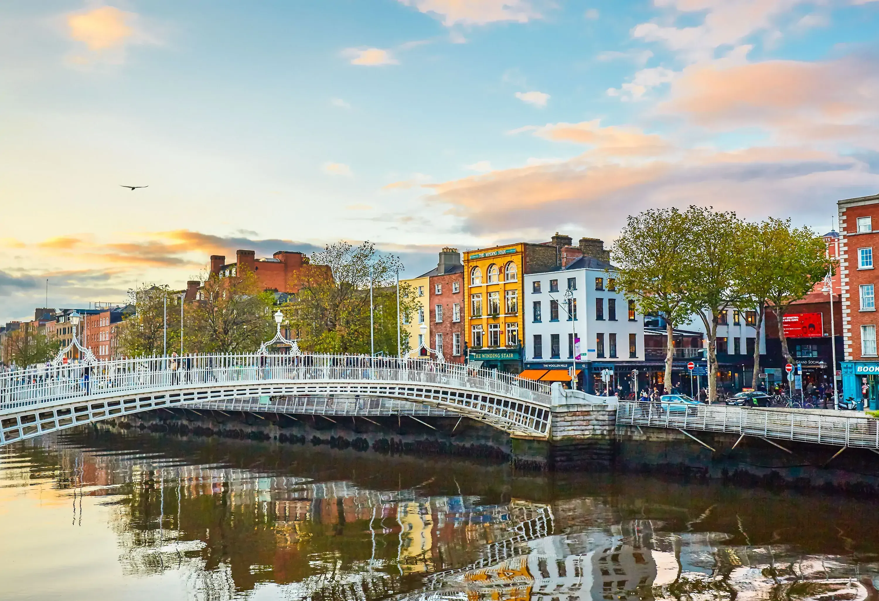 An iron footbridge across a river with colourful buildings in the background.