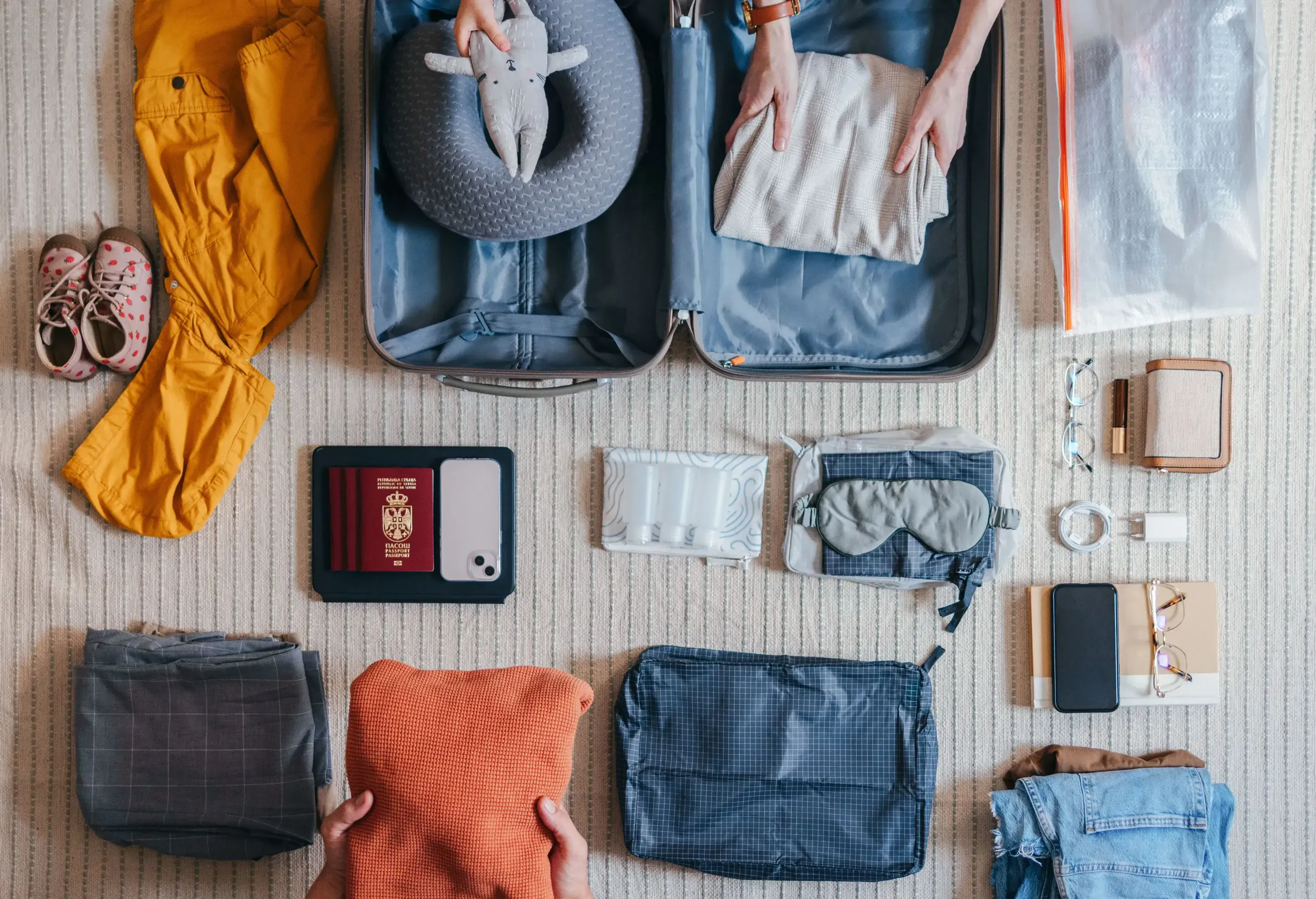Overhead view of persons' hands packing a suitcase with clothes, with various travel items arranged around it.