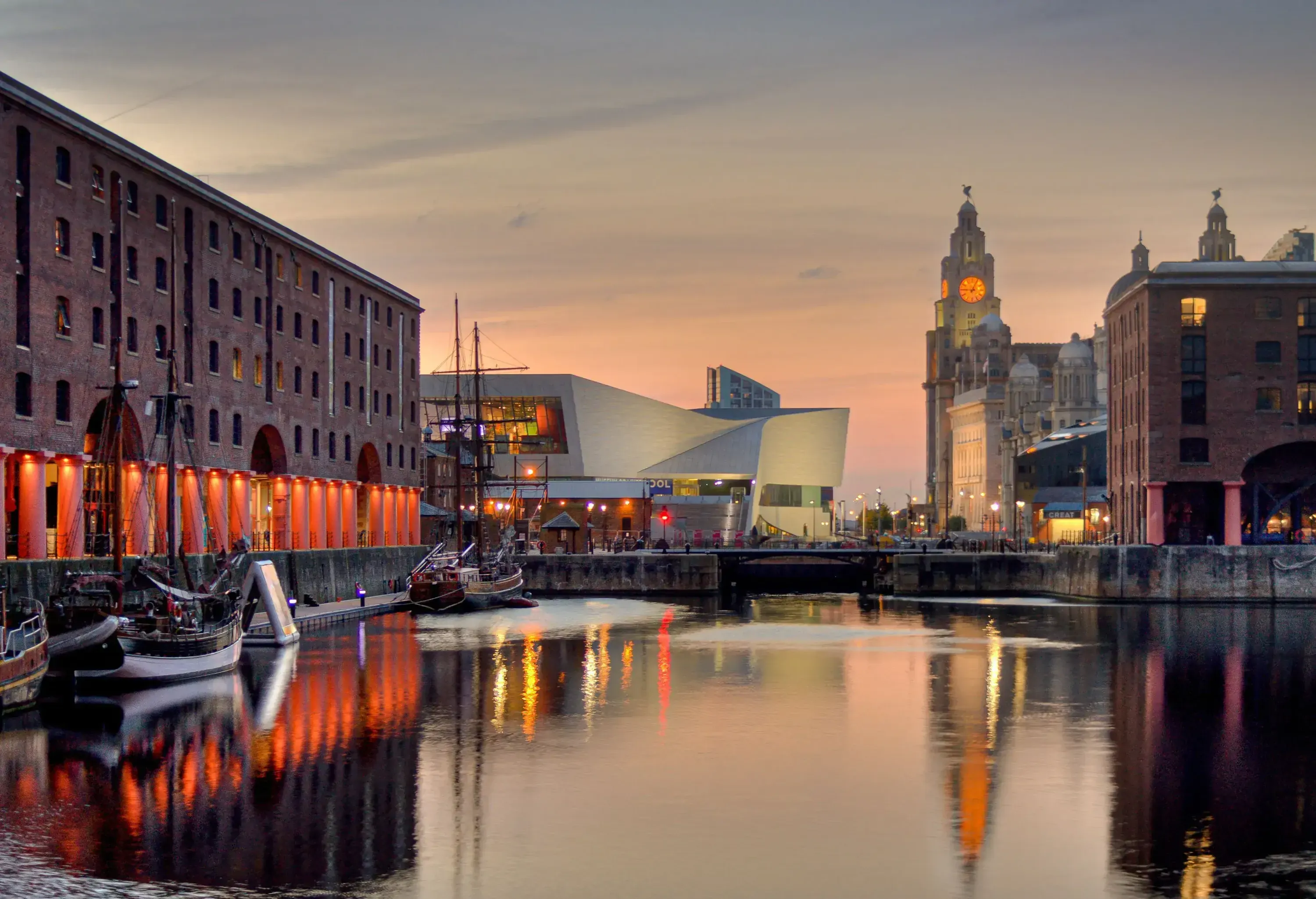A dock adjacent to a bridge surrounded by buildings with a view of a lit clock tower in the distance reflected on the water's surface.