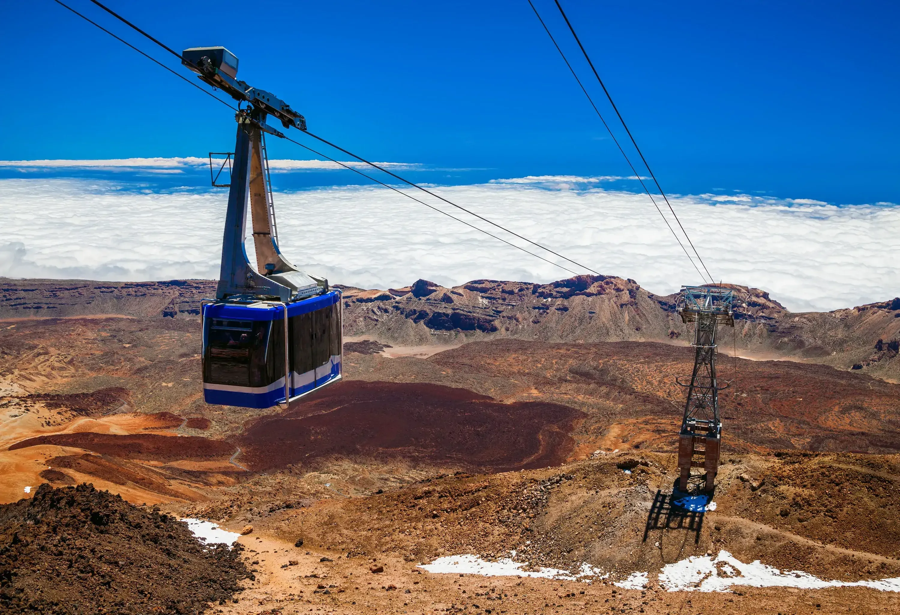 Cable cars travelling over a volcanic terrain with a sea of white clouds and the blue sky in the background.