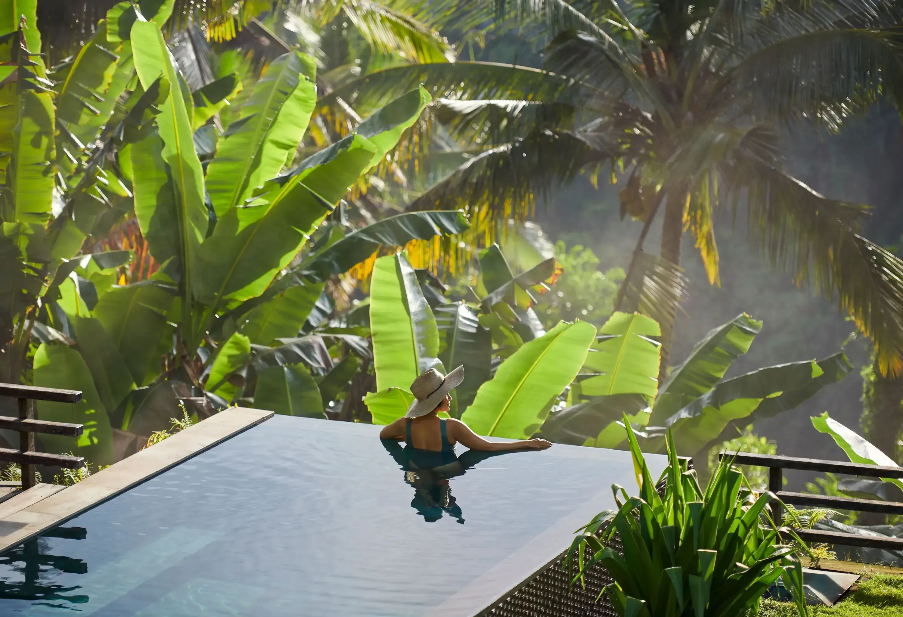 Woman wearing a hat relaxing in an infinity pool, overlooking a lush, green tropical environment.