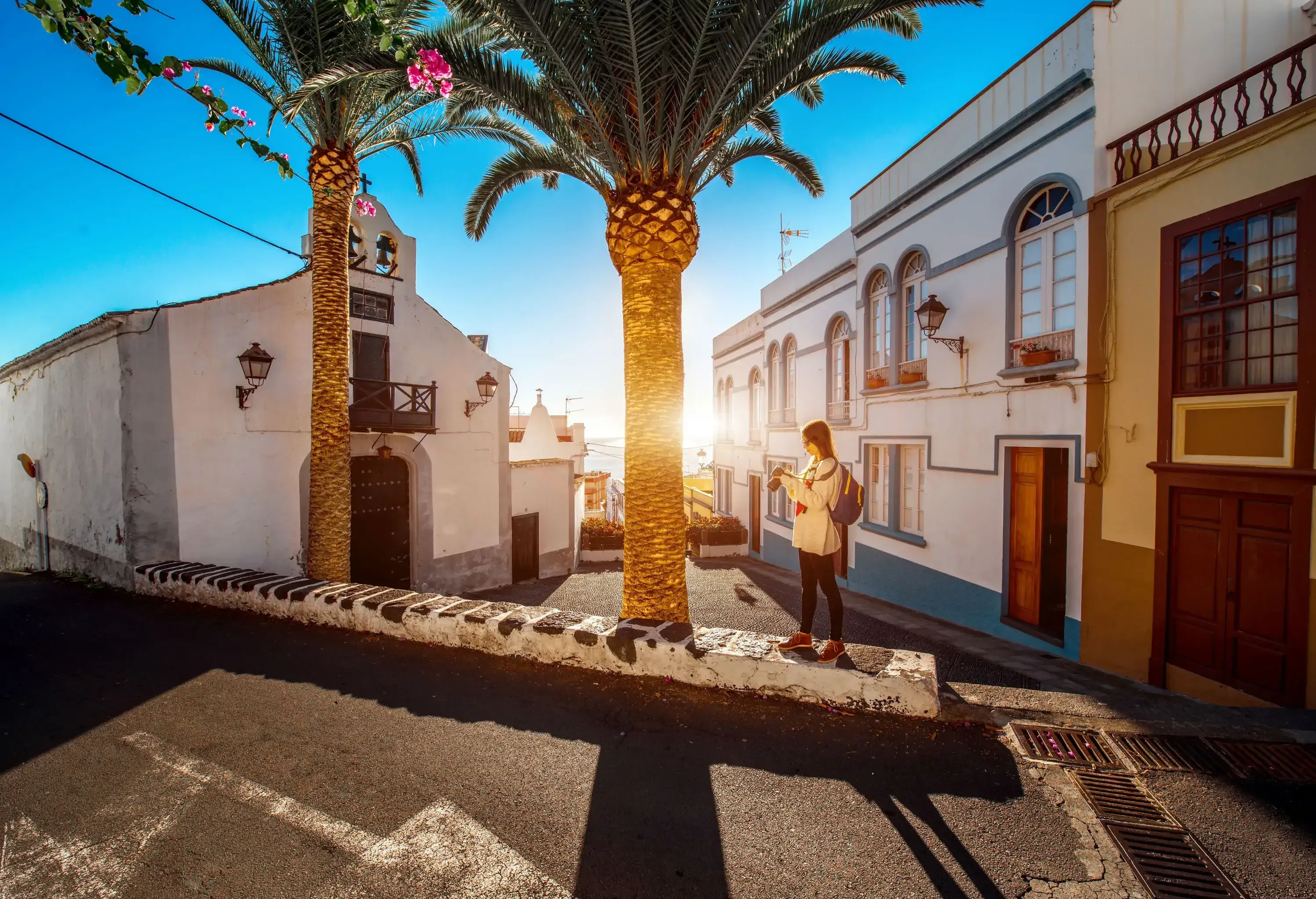 A woman standing on the curb of an old town street with trees blocking the sunlight.