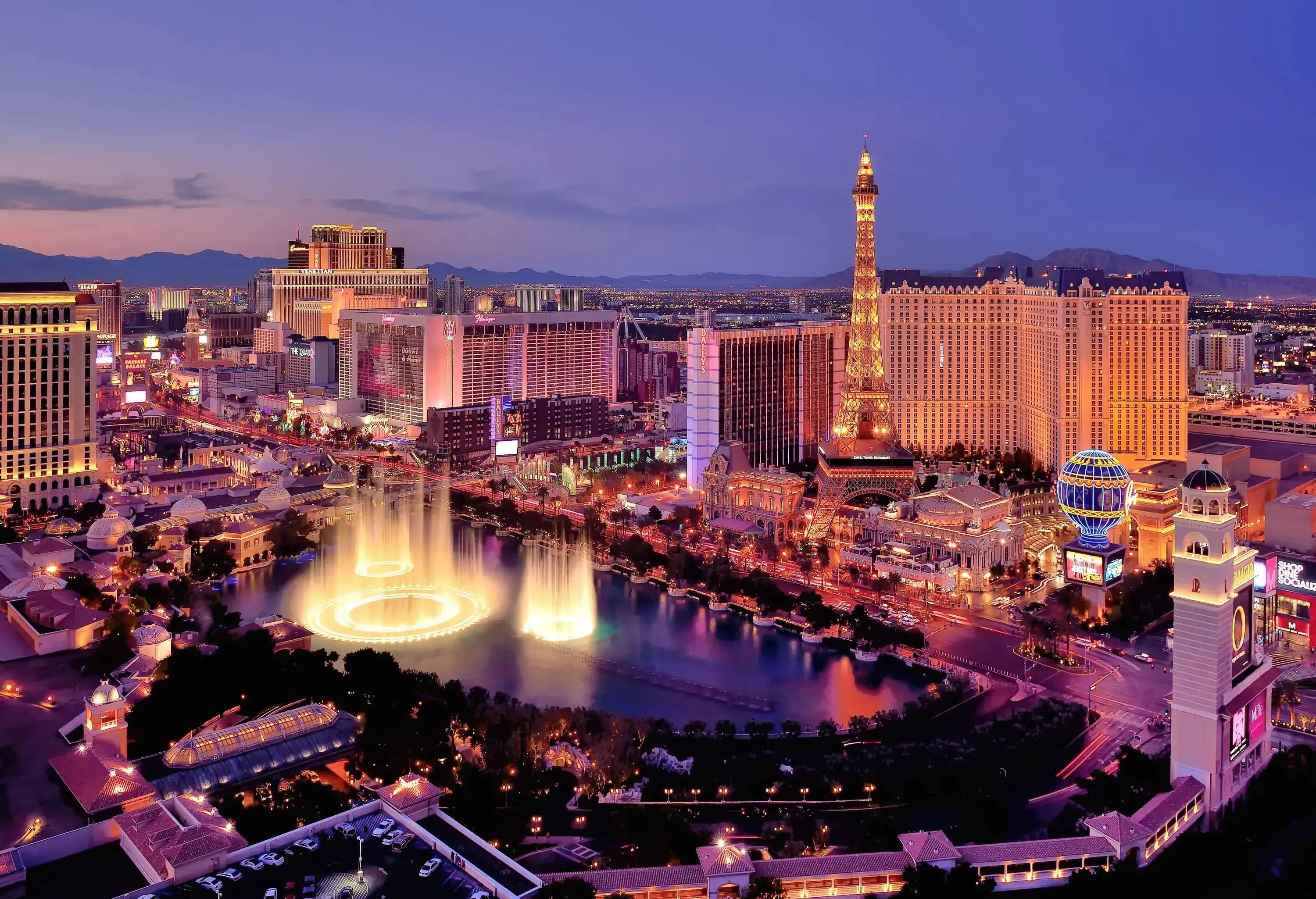 A tall replica of the iconic Eiffel Tower stands near a beautiful illuminated fountain amidst the tall buildings.