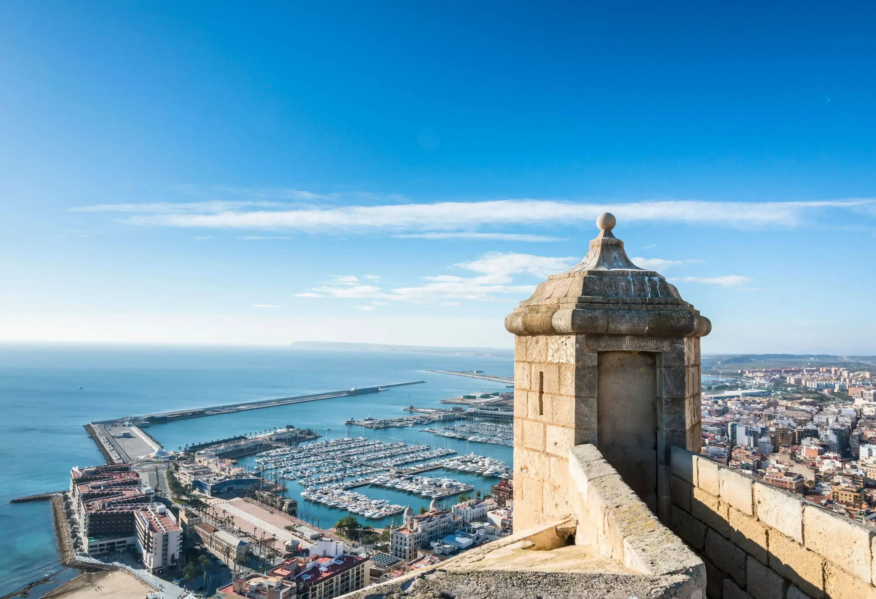 An ancient stone watchtower stands sentinel, surveying the bustling harbour below, where docked boats gently bob alongside the vibrant coastal town.