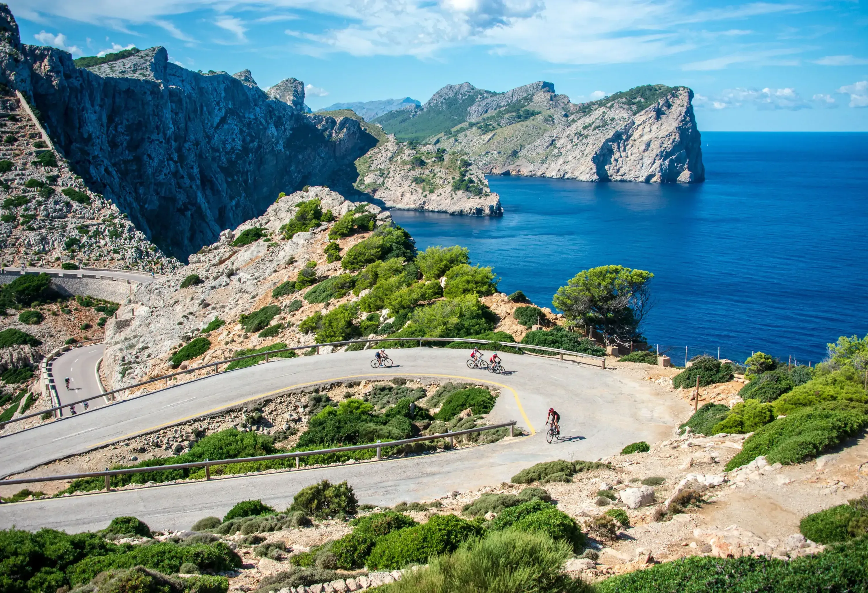 A winding coastal road snakes along dramatic cliffs overlooking a deep blue sea, with several cyclists visible on the road.